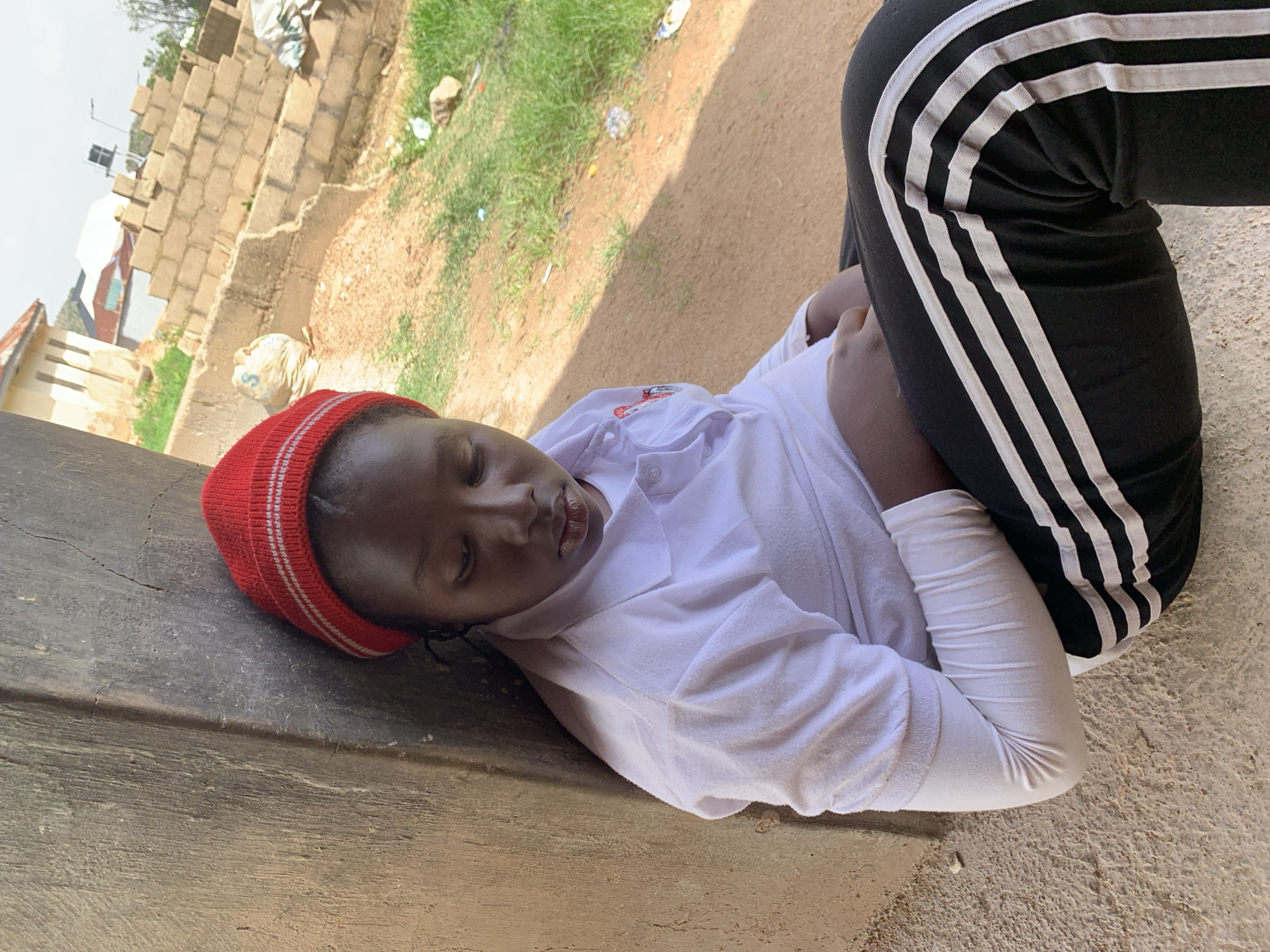 A young person wearing a red knitted beanie, a white collared shirt, and black pants with white stripes is sitting with their eyes closed, leaning against a textured, rough-surfaced wall. Their head is tilted down, and their hands are resting in their lap. The environment appears to be outdoors with dry, sandy ground and patches of green grass. In the background, there is a partially constructed building made of concrete blocks, some sacks, and what looks like debris. The overall scene suggests an urban or semi-urban setting in a warm climate. This location is identified as the city of Jos, Nigeria.