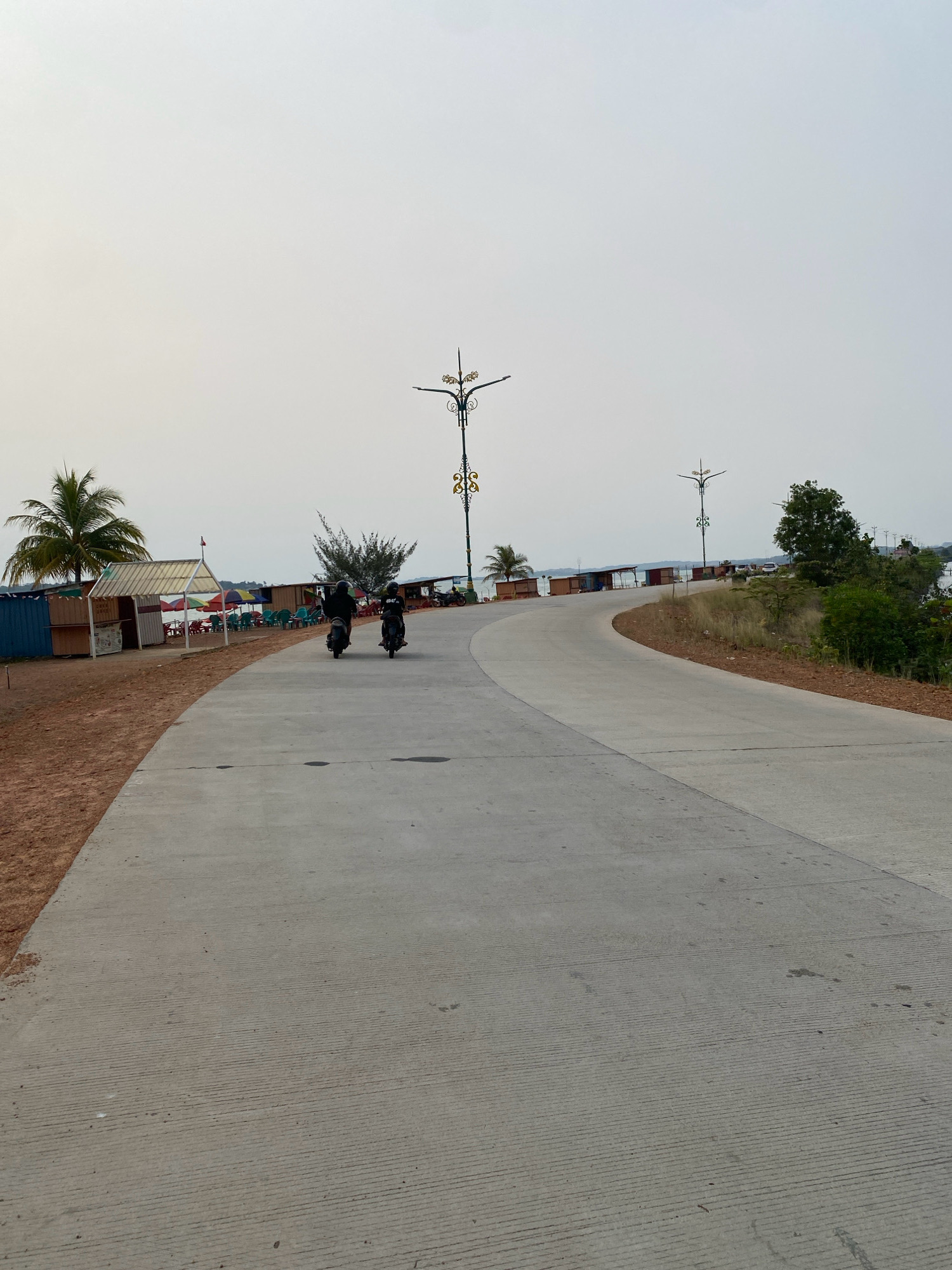 The image depicts a concrete road winding through a coastal area in Tanjung Pinang, Indonesia. On the left side of the road, a sandy beach is visible, lined with small wooden structures that appear to be shops or food stalls, equipped with colorful umbrellas and chairs. A palm tree stands prominently near one of these structures. Further down the road, two individuals are riding scooters, heading away from the camera. The road itself is marked with parallel lines, suggesting it is designed for vehicles. In the background, the sky is overcast, and a body of water can be seen beyond the buildings. Decorative streetlights with intricate metalwork are positioned along the road. The overall scene suggests a relaxed, possibly tourist-oriented, environment by the sea.