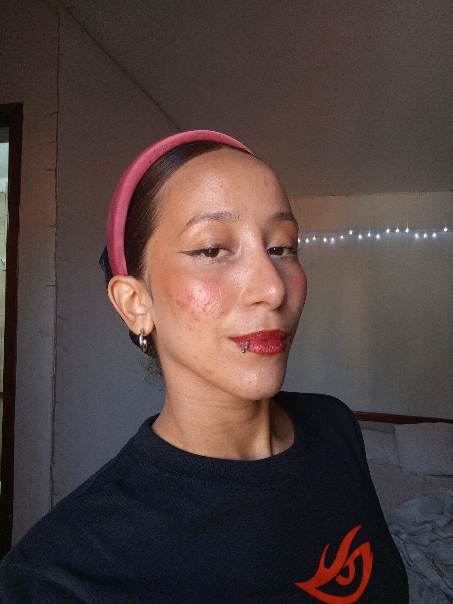 A medium indoor shot depicts a woman with fair to medium skin wearing a pink headband, black eyeliner, red lipstick, and a black t-shirt with a red design. She has a piercing on her lower lip and is wearing hoop earrings. Her cheeks appear flushed or have makeup applied. Behind her is a wall with string lights and a partially visible bed. The location of the photo appears to be indoors, but the city of Porlamar, Venezuela, cannot be identified from the image.