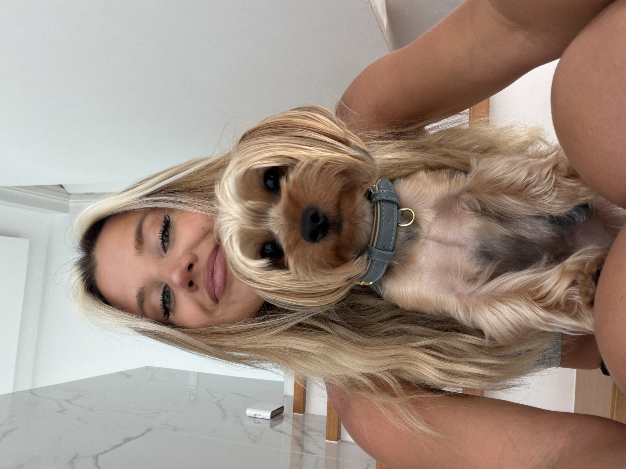 A young Caucasian woman with blonde hair is taking a selfie with a small Yorkshire Terrier. The woman is wearing a grey top, and the dog is wearing a grey collar. They are sitting on stairs, likely in a modern building with white walls and a marble accent on the left. The background includes a white railing and a glimpse of stairs. The lighting suggests an indoor environment, possibly a home or a hotel, with natural light. The woman is smiling, and the dog is looking at the camera. There is no indication of the city of Thep Krasatti, Thailand in the image.