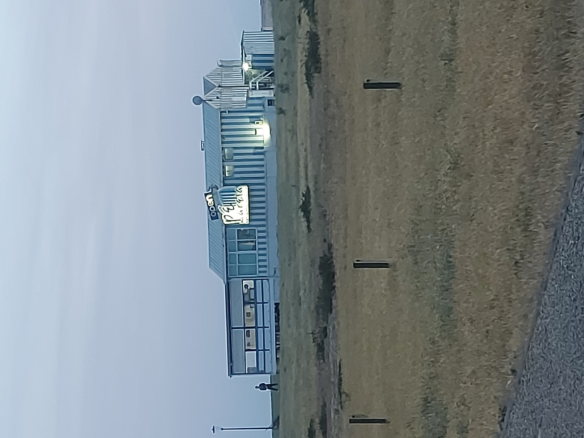 A long-shot photograph of the "Pãdaria Mareia" bakery in Figueira da Foz, Portugal, appears to be taken from across a grassy field. The building has a vertically striped exterior with the bakery's sign, which reads "Pãdaria Mareia", illuminated and displaying "Open." A person is walking in front of the building. Utility poles are also visible.