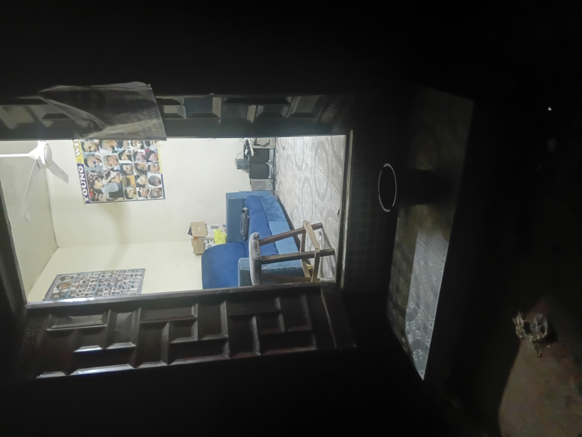 The image displays an interior space viewed through a partially open, dark wooden doorway with decorative paneling, from a darker foreground area. The scene is located in the city of Jalingo, Nigeria.
The foreground, outside the doorway, is dark brown/black, showing a small, dark bucket with a light-colored rim on a slightly elevated surface to the right. To the left, a piece of crumpled, light-colored material lies on the ground. A white chalk circle is visible on the ground surface immediately in front of the bucket.
The interior space, illuminated by unseen light sources, features light-colored (cream/off-white) walls and a white ceiling with a white ceiling fan mounted centrally. The floor is covered with light-colored tiles featuring a repeating circular or floral pattern.
Furniture includes a blue upholstered sofa, possibly L-shaped, positioned against the back and right walls. A wooden stool with a light-colored, possibly grey, padded cushion is placed in front of the sofa. In the far right corner, a stand holds electronic equipment, including speakers and what appears to be a receiver or media player. Stacked cardboard boxes are visible on the floor in the background, behind the sofa.
Two large posters adorn the walls. On the left wall, a rectangular poster displays a grid of multiple small photographs, likely portraits. On the back wall, a larger, horizontal poster features the text "YOUNG - WIN" and a grid of distinct male hairstyles and portraits, strongly indicating a barbershop or hair salon setting. A piece of light-colored fabric or paper is draped over the right door frame, hanging into the interior space.
No individuals are present or visible in the scene.