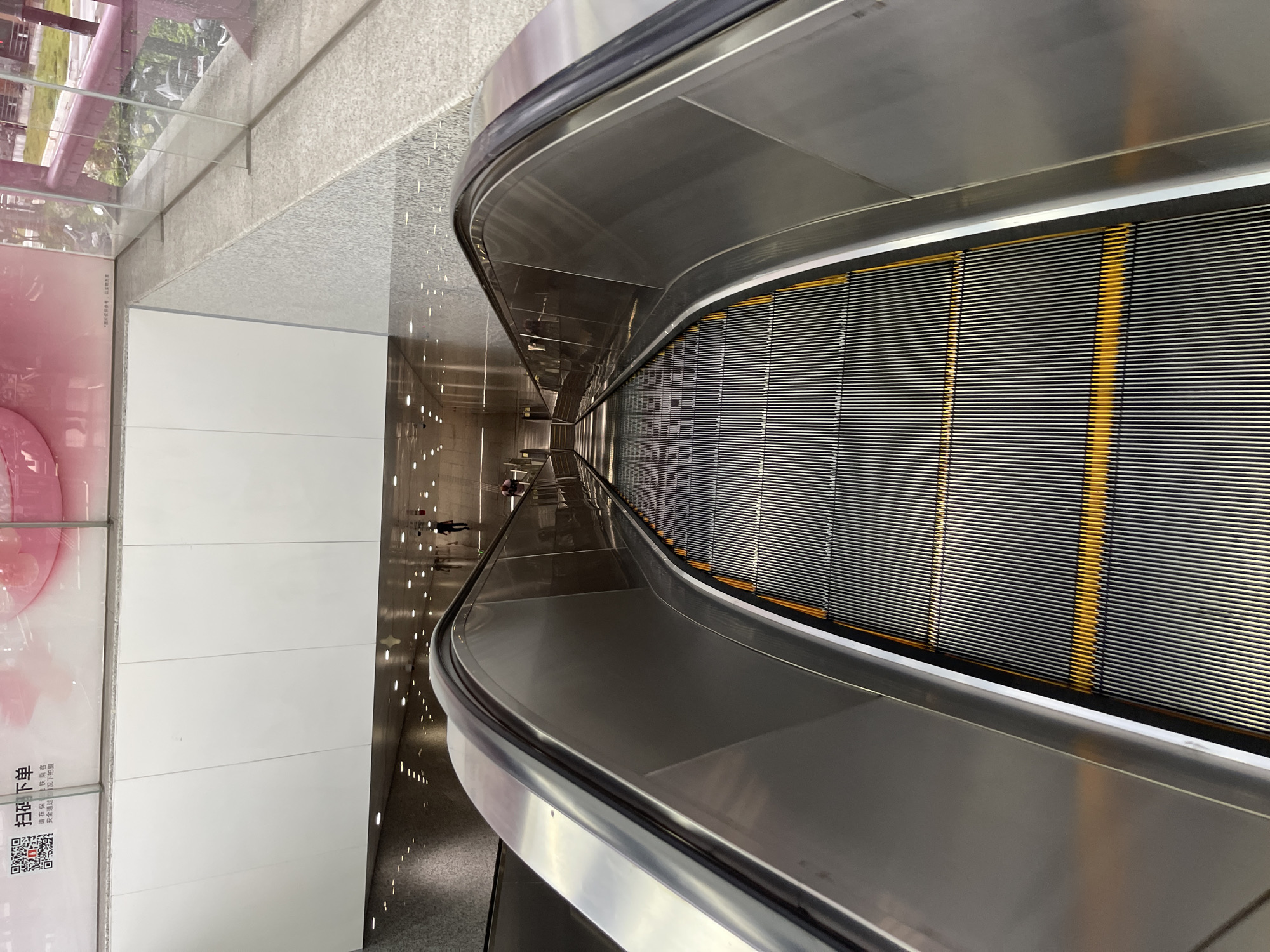 This media file shows a view from the top of a moving escalator, looking downwards. The escalator has metal steps with yellow safety lines and a brushed metal handrail. The surrounding environment appears to be an indoor public space, possibly a shopping mall or transportation hub, given the polished floors, modern architecture, and soft lighting. To the left of the escalator, a large white wall is visible, featuring a pink advertisement with some Chinese text and a QR code. Further down the hall, several people can be seen walking. The scene is clean and well-lit, suggesting it is daytime. The overall impression is one of a modern and efficient urban environment. The text on the advertisement reads "扫码下载" which translates to "Scan code to download".
