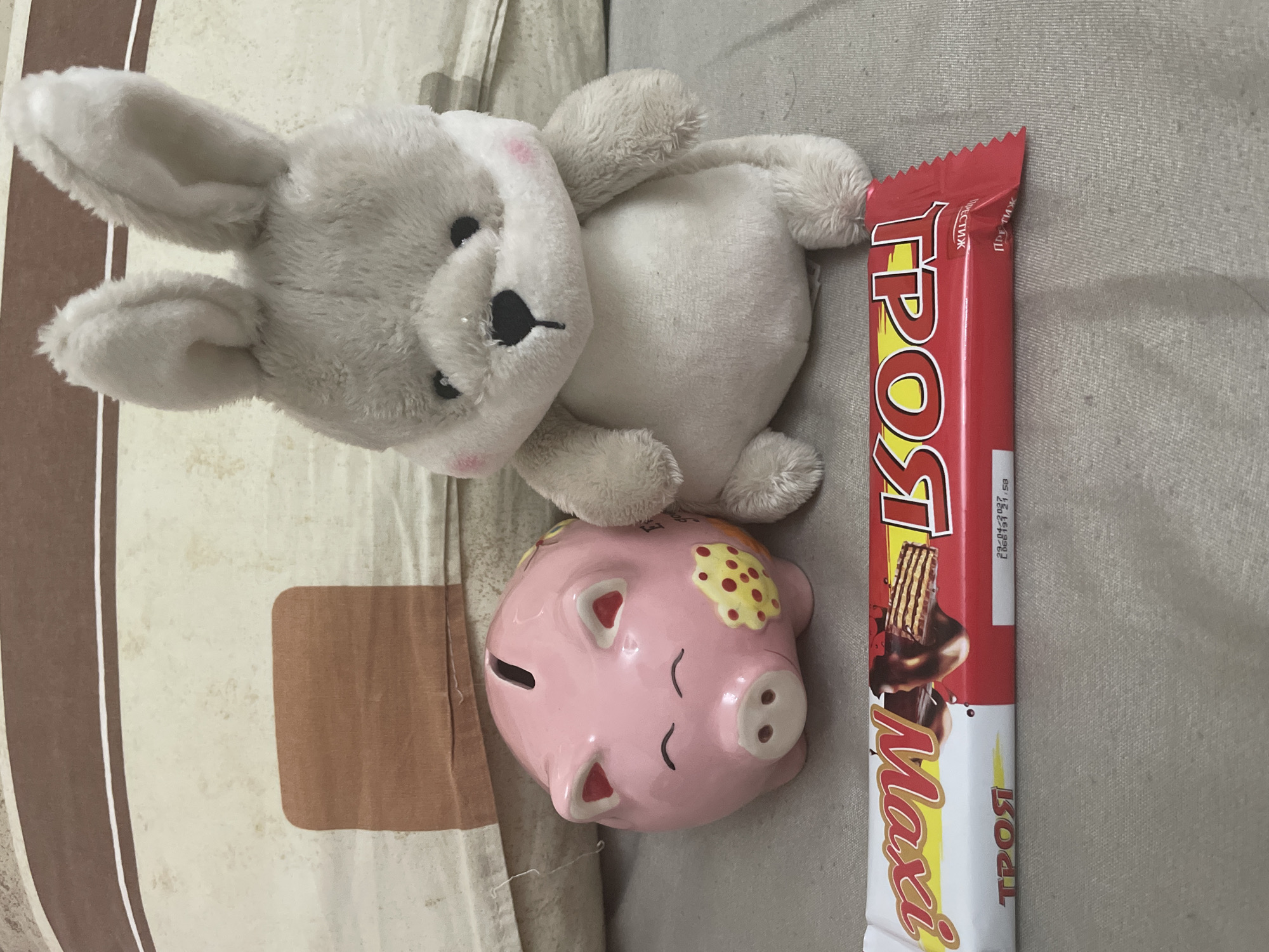 This image captures an indoor scene in the city of Ihtiman, Bulgaria. In the foreground, a red and white packaged chocolate bar lies horizontally on a light grey textured fabric surface. The white section of the wrapper displays "Maxi" (upside down) and "ТРОЯ" (Troya in Cyrillic), while the red section shows a chocolate-coated wafer image, the word "ТРОЯ" (Troya), and product details including "29/04/2027" and "L066191 21:58".
In the midground, a pink ceramic piggy bank is situated to the left of a light grey plush rabbit toy. The piggy bank features a coin slot on top, closed black eyes, a snout with nostrils, and yellow polka-dot decals on its side. The plush rabbit, characterized by its upright posture, black eyes, black nose, and pink blushed cheeks, sits to the right of the piggy bank.
The background consists of a large striped pillow or cushion. It exhibits horizontal bands of brown, a light cream or beige color with subtle discolored spots, and a thin white stripe. A solid brown rectangular fabric patch is visible on the light cream section of the pillow.