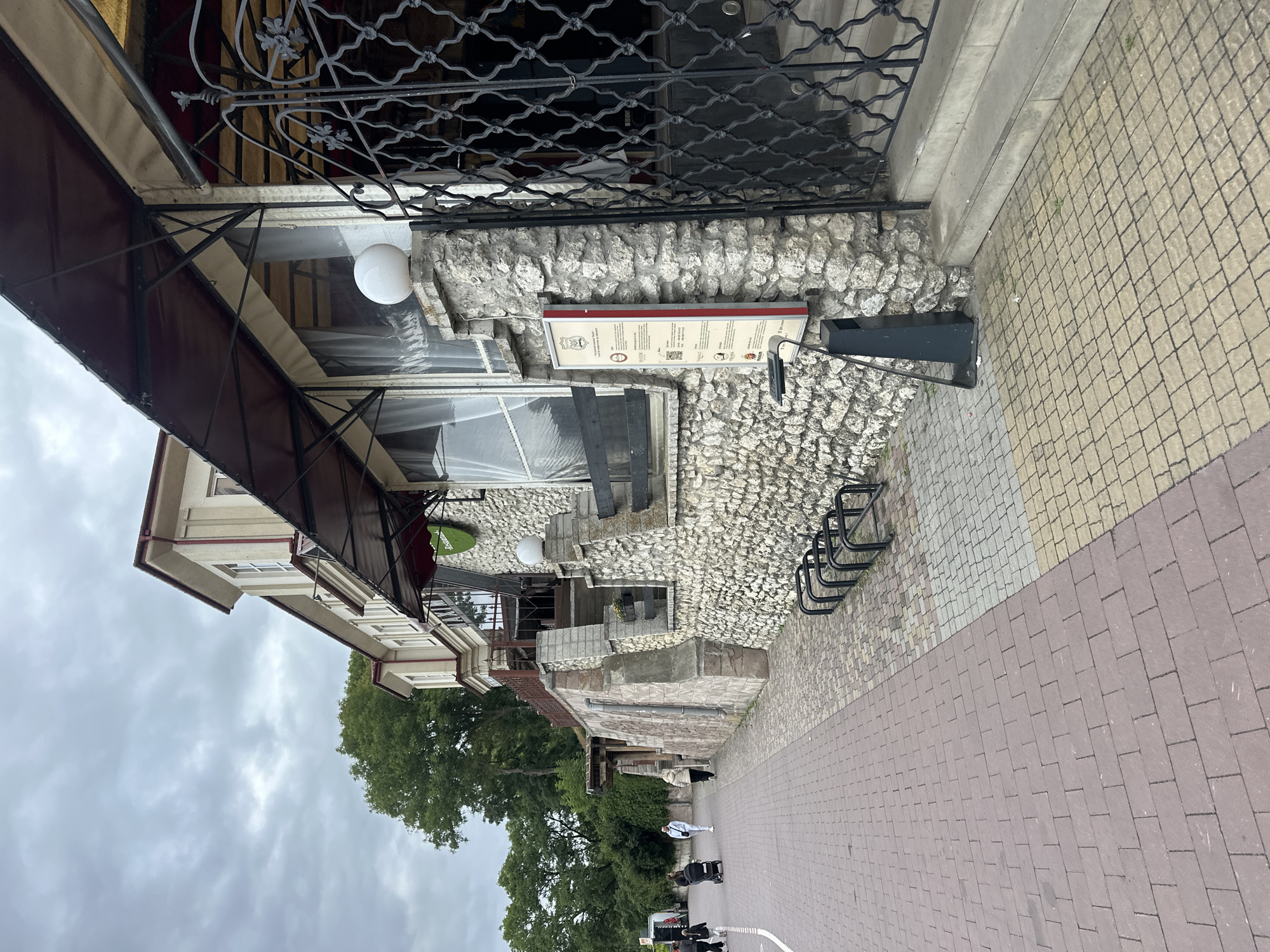 A stone-clad building with a pitched roof and awnings is seen from a street-level perspective. The building has multiple windows covered with translucent curtains and ornate metalwork at the entrance. A green sign with white lettering is visible on the building's facade. In front of the building, there are a few bike racks and a trash receptacle. A sidewalk made of brick pavers runs along the side of the building, with people walking and a stroller present. The sky is overcast. This scene is likely located near the Ternopil Lake in Ternopil, Ukraine, given the building's design and proximity to a waterfront area often featuring walking paths and recreational facilities.