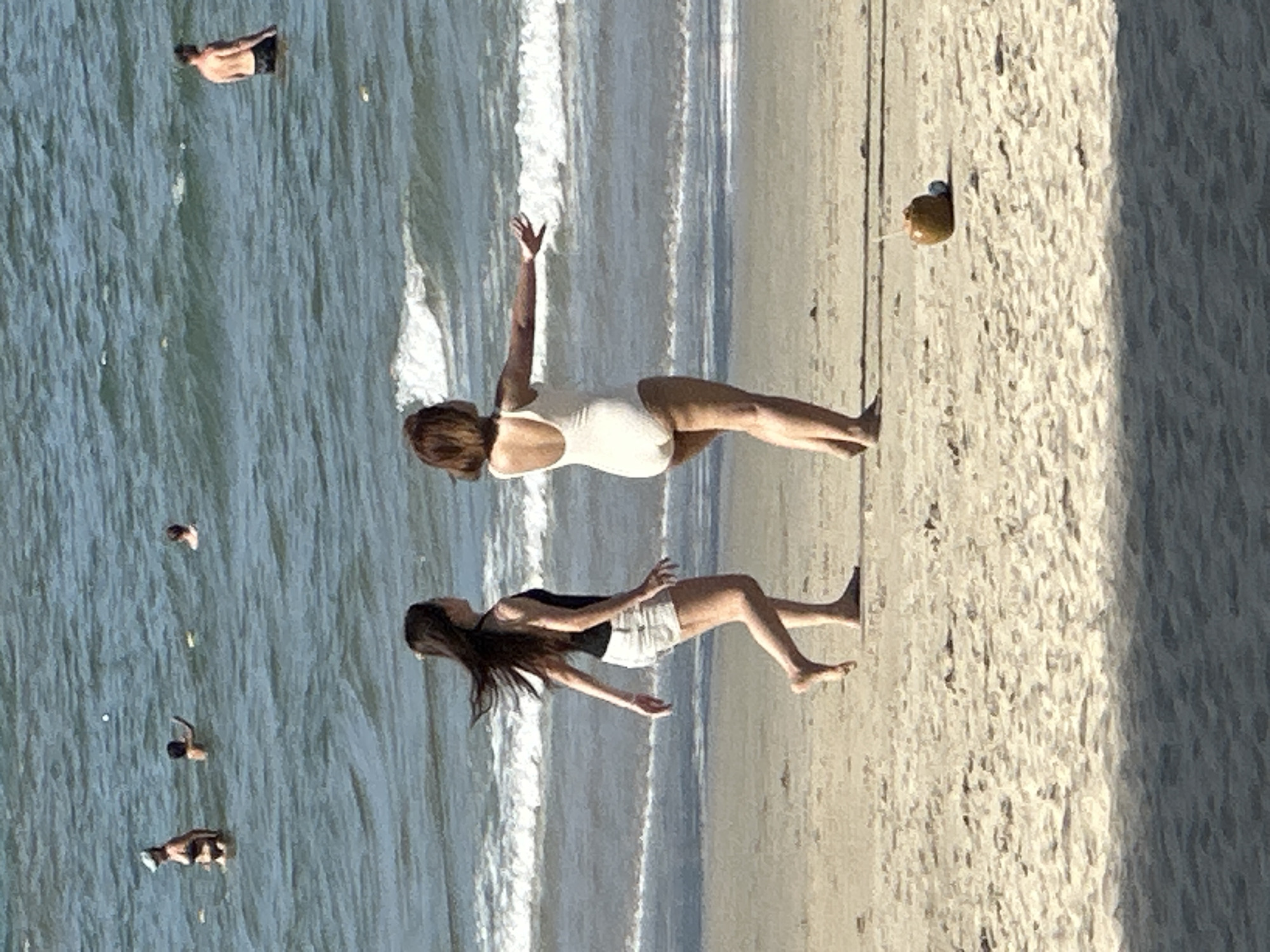 On a sandy beach in Đà Nẵng, Vietnam, two women are walking near the water's edge. The woman on the left wears a black tank top and white shorts, with her long dark hair blowing in the wind. The woman on the right is in a white one-piece swimsuit and appears to be gesturing with her outstretched arm. In the foreground on the sand, a coconut with a straw rests next to a small blue object. In the background, several people are in the ocean, with waves breaking on the shore. The water is a blue-green color under a bright sky.