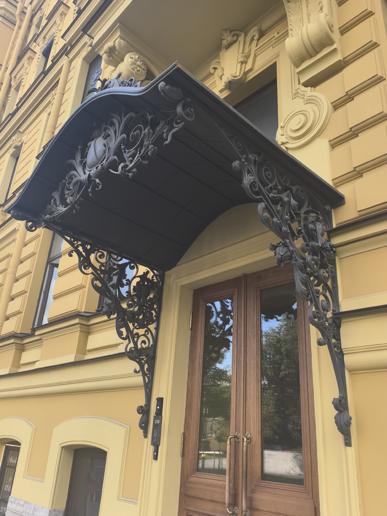 The image shows the exterior of a building with a yellow facade, characteristic of architecture found in Saint Petersburg, Russia. A black ornate metal canopy extends over a double wooden door. The canopy features intricate scrollwork, floral motifs, and a central shield-like emblem. Decorative plasterwork adorns the building's facade around the canopy and windows, typical of historic European architecture.
The double doors are made of wood with glass panels that reflect the surrounding environment, including trees and the sky, suggesting a daytime setting on a clear day. A modern intercom system is mounted on the wall next to the door. Below the canopy, there are two arched doorways, one of which is partially visible and appears to lead to a shop or another entrance.
The overall impression is one of historical grandeur and architectural detail. The lighting indicates it is daytime, likely with bright sunshine given the reflections. The building's style and the presence of ornate metalwork are strong indicators of a European, specifically Russian, urban setting. There is no visible text in the image other than what might be on the intercom display.