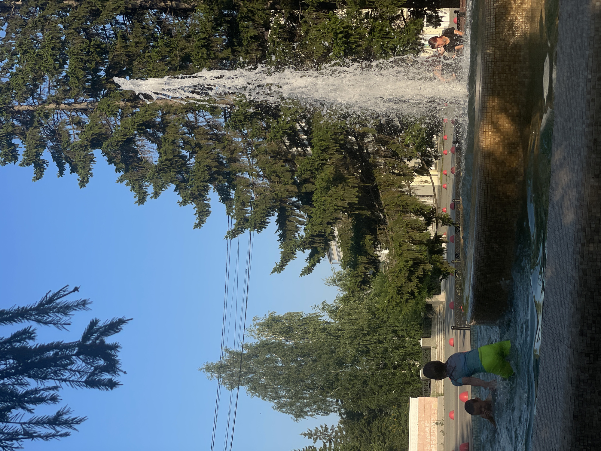 In a city park under a clear blue sky, two children are playing in a fountain. The fountain features a tall, powerful jet of water. On the left, a younger child in a blue shirt and green shorts splashes in the water, with their face partially submerged. Beside them, another child is also playing in the water. In the background, several large evergreen trees and a birch tree are visible, along with a building and red dome-shaped objects scattered on the ground. Another person is partially visible behind the water jet, appearing to be in the fountain as well. The fountain has a tiled edge.
