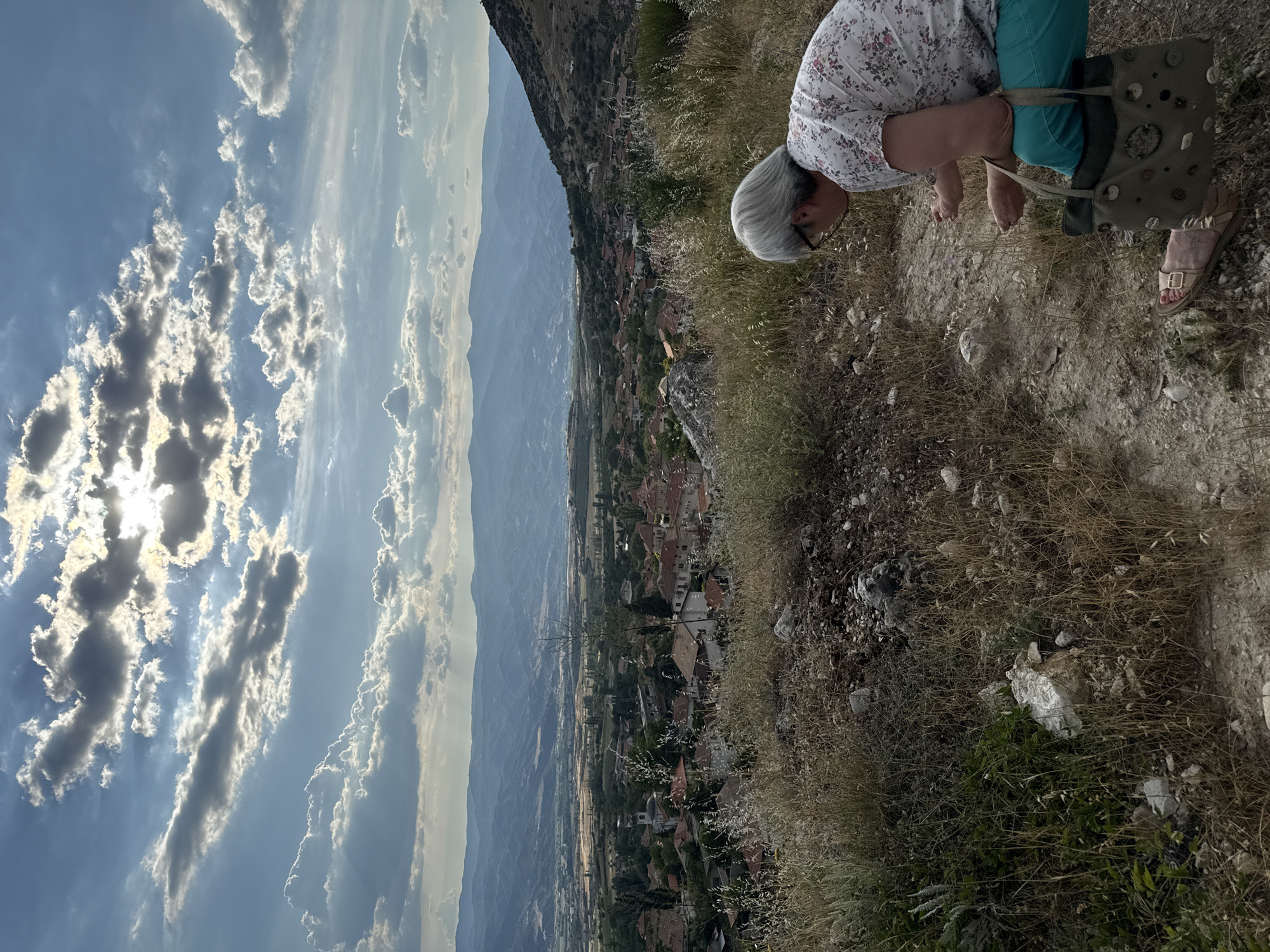 An elderly woman with short gray hair and glasses is crouching down, examining something on the ground. She is wearing a white shirt with a floral pattern and teal pants. A large, olive-green tote bag with various decorative circular embellishments rests beside her. The woman is positioned in the foreground, on a dry, grassy hillside dotted with rocks and sparse vegetation.
Behind her, a panoramic view unfolds of the town of Ilindentsi, Bulgaria. The town is nestled in a valley, characterized by numerous terracotta-tiled roofs and scattered trees. Further in the distance, rolling mountains recede into a hazy blue horizon. The sky above is dramatic, with a mix of dark, textured clouds and bright rays of sunlight breaking through, creating a sense of diffused light. The overall scene captures a moment of observation amidst a scenic, somewhat rugged landscape.