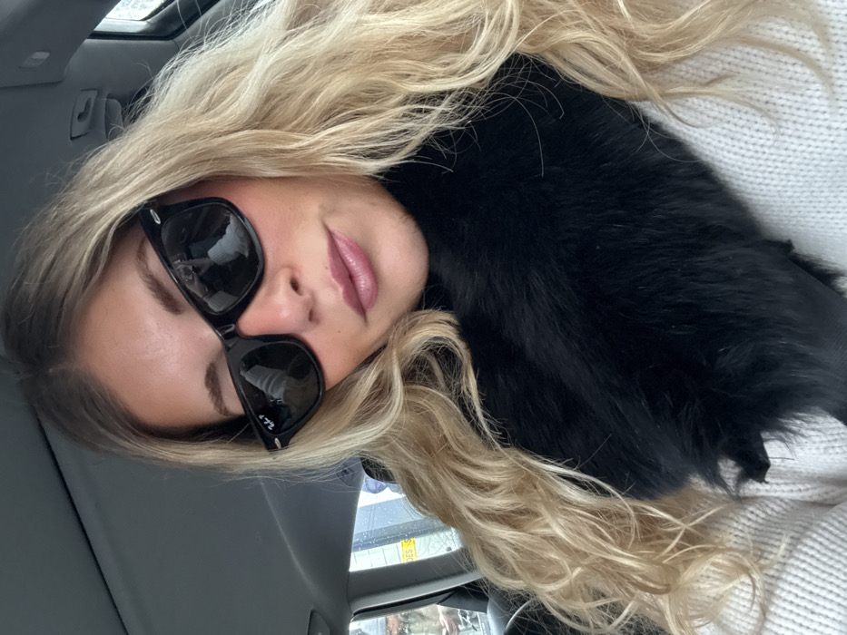 A female subject with long, wavy blonde hair and a tanned complexion is depicted from the chest up, looking directly at the viewer. She is wearing black Ray-Ban Wayfarer sunglasses, a black fur collar or scarf, and a cream-colored knitted sweater. Her lips are made up with a pink/mauve lipstick. The subject is seated within a vehicle, with the interior ceiling visible above her head. Through the vehicle's window on the left, a blurred urban street scene is visible, featuring a light-colored building and a white street sign. The sign displays dark text, partially legible as "DES S." followed by further indistinct letters. This lettering is consistent with street signage for Rue des Saints-Pères, placing the location in Paris, France.
