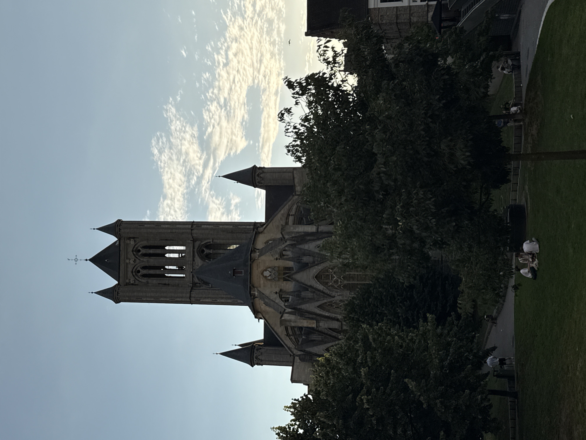 This is a photograph of Saint Nicholas' Church in Ghent, Belgium, taken from a park area in front of it. The church, a prominent landmark in the city, is a Gothic-style cathedral with a tall central tower and two smaller flanking towers. Its facade features intricate stonework and arched windows. In the foreground, several people are seated on the grass, seemingly enjoying a leisurely afternoon. Lush green trees partially obscure the lower sections of the church and extend across the foreground, framing the scene. The sky above is a pale blue with scattered clouds, suggesting it is either early morning or late afternoon. The overall atmosphere of the image is peaceful and serene.