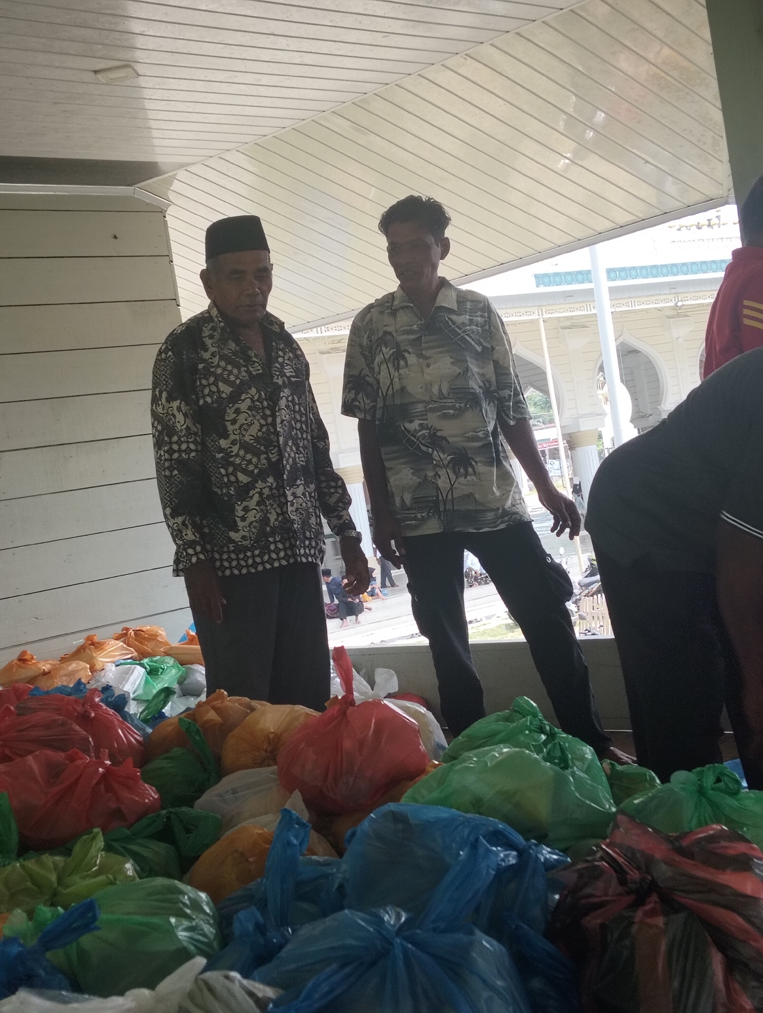 The image depicts a scene in Grong-Grong, Indonesia, featuring two adult males standing near a large collection of colorful plastic bags, likely containing food or donations. The man on the left wears a traditional peci hat and a patterned shirt, while the man on the right is dressed in a short-sleeved, patterned shirt. Both are looking towards the right of the frame. In the background, a building with arched windows and a pale facade suggests a public or religious structure. The foreground is dominated by the numerous plastic bags, which are arranged in piles and vary in color, including red, green, blue, orange, and white. The overall lighting suggests it is daytime, possibly outdoors or in a covered area with natural light.