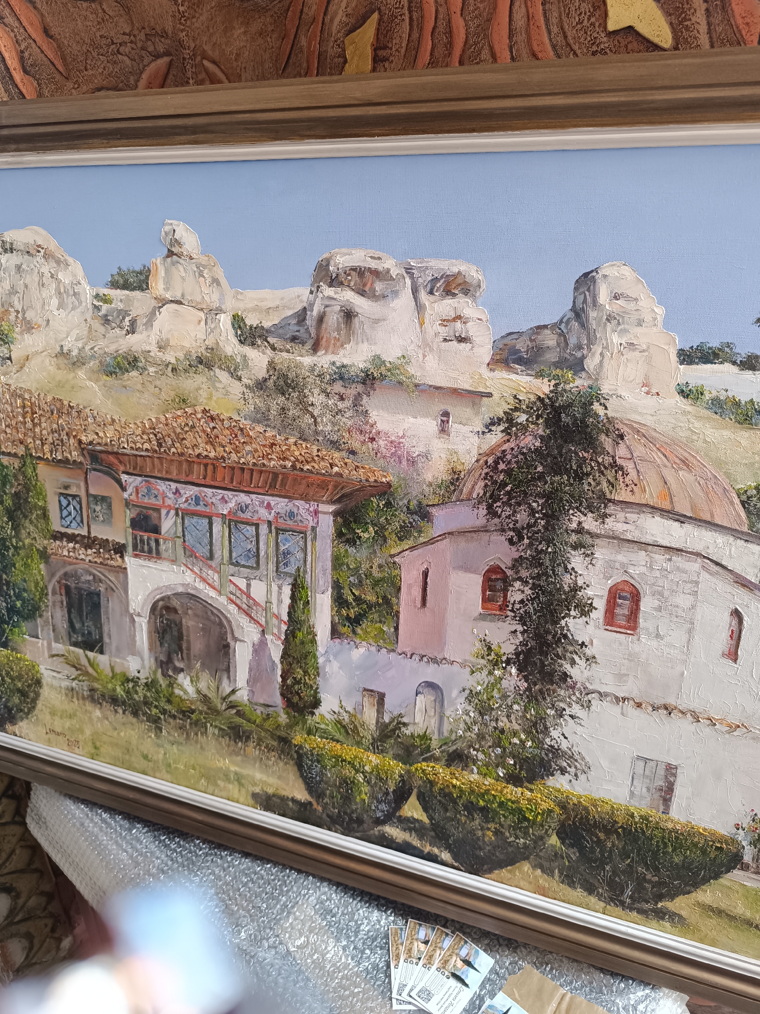 The image shows a framed oil painting of the Bakhchisaray Palace, located in the city of Ukromne, Ukraine. The palace complex is depicted with its distinctive architecture, including the tile-roofed buildings with arched walkways and ornate decorations. The adjacent mosque with its dome is also visible. In the background, large rock formations rise above the palace buildings. The sky is a clear, pale blue. The painting is encased in a decorative frame, and partially obscured by bubble wrap, cards and flooring beneath.