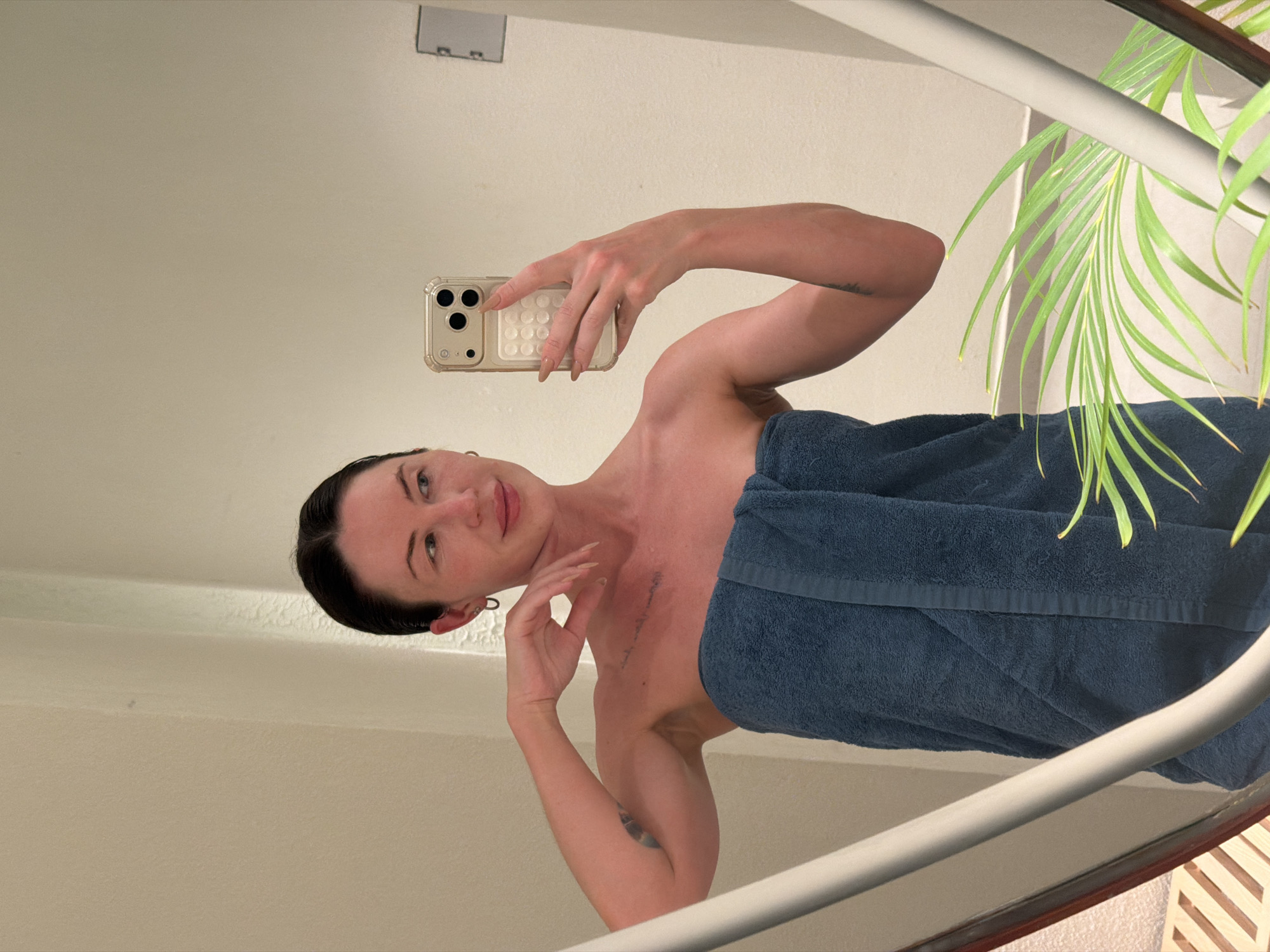 In a brightly lit bathroom, a woman takes a mirror selfie. She is wearing a blue towel wrapped around her body and has her dark hair pulled back. Her left arm is bent at the elbow, with her hand resting on her head, and she is looking at her reflection with a slight smile. In her right hand, she holds a smartphone, also angled towards the mirror. The bathroom features a light-colored wall, a large mirror with a thin frame, and a lush green plant in the foreground. A partial view of wooden shelves is visible on the left side of the frame. There are no visible indications of the location being in Dong Ninh Hoa Ward, Vietnam.