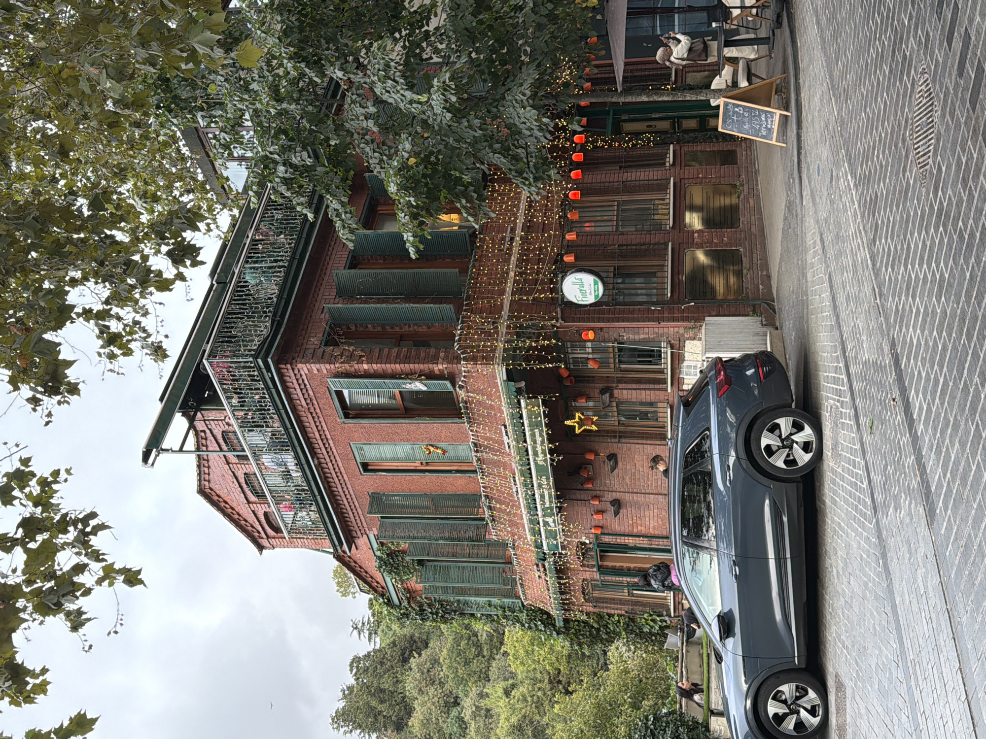 The image captures a brick building in Üsküdar, Turkey, likely a restaurant or cafe, adorned with string lights and decorative elements. The building has multiple floors, with open windows and shutters. A balcony with decorative railings is visible on the upper floor, and people are seated there. In front of the building, a grey Audi e-tron electric car is parked on a cobblestone street. Several people are standing and sitting outside the building, some near the car, and others at tables under an umbrella. A chalkboard sign with text is propped up on the sidewalk. Lush greenery, including trees and ivy-covered walls, surrounds the building, suggesting a picturesque setting within the city. The sky is overcast.