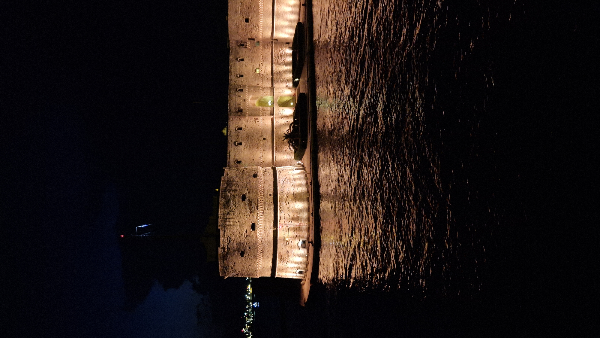 A castle, illuminated by warm lights, stands on the edge of a body of water at night. The castle is made of stone and has a circular structure with battlements. The lights create reflections on the water's surface, which appears to be slightly rippled. In the background, lights from buildings can be seen in the distance, suggesting an urban environment. A flagpole with a red and blue light is visible atop the castle. The sky is dark and cloudy. This is likely the Aragonese Castle in Taranto, Italy, given its prominent location on the sea and its distinctive architecture.