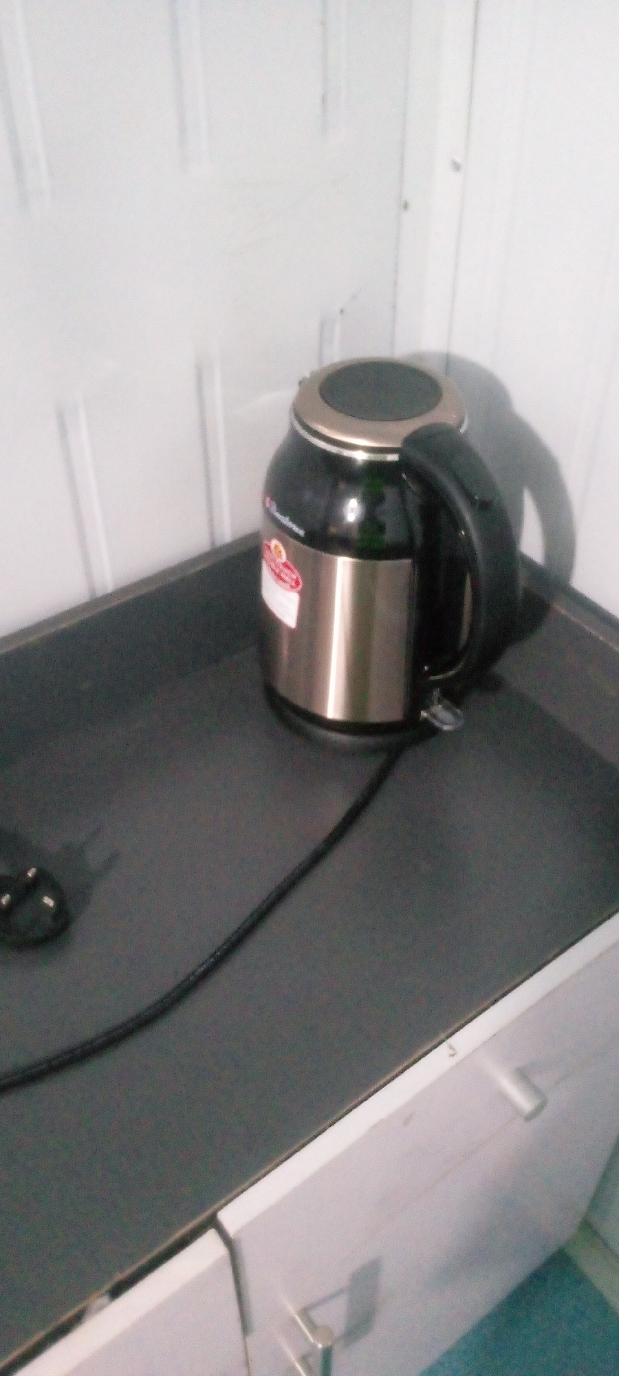 The image depicts an electric kettle placed on a dark countertop. The kettle is black and silver, with a black handle. A power cord extends from the kettle and lies across the countertop. The countertop appears to be part of a kitchen counter or a piece of furniture. In the background, there is a white, ribbed wall, possibly part of a cupboard or cabinet. The lighting is somewhat dim, casting shadows. The scene is indoors, and the specific location in Indo, Nigeria cannot be determined from the image alone. There are no people or any discernible actions occurring in the image.