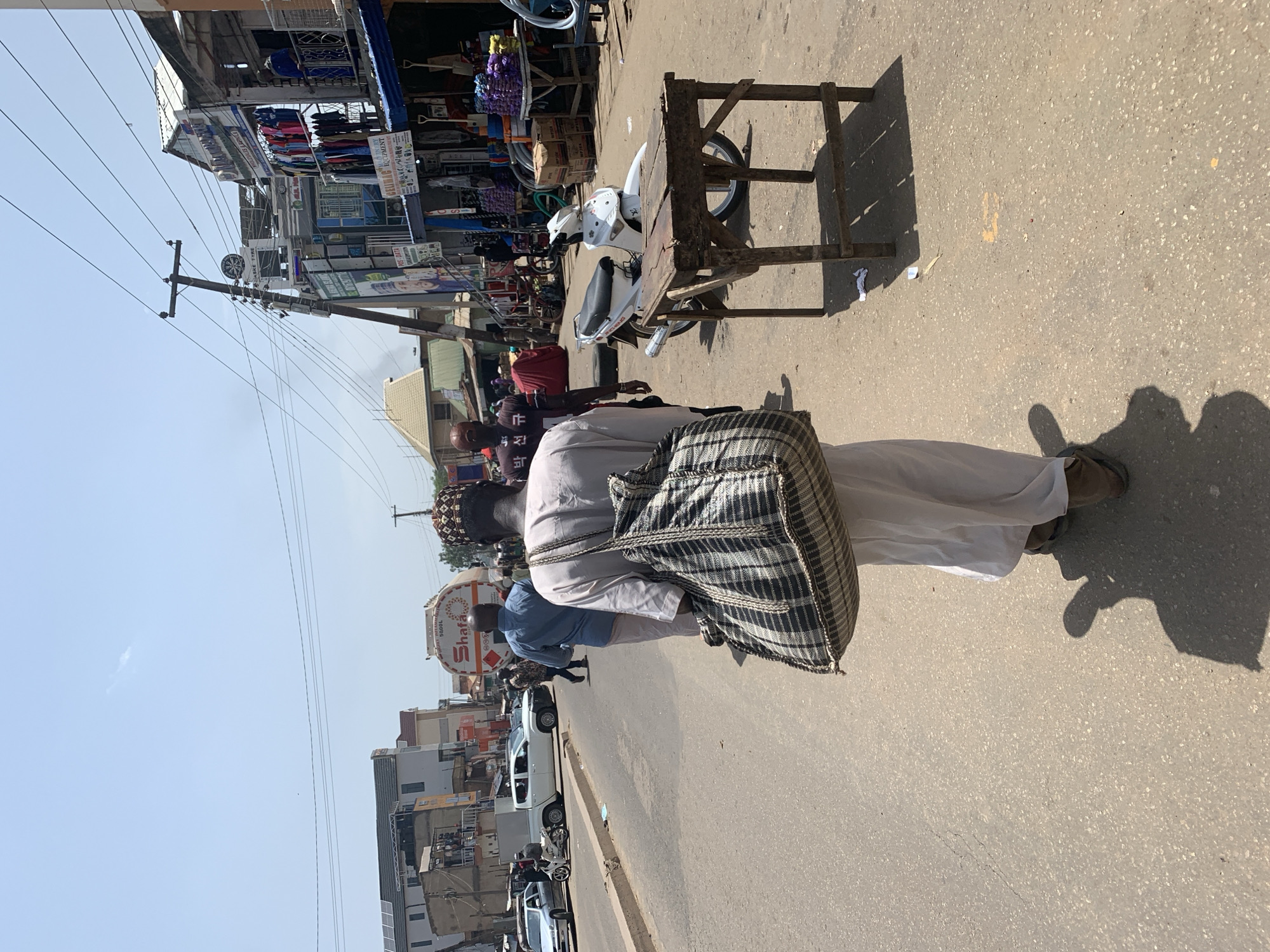 The image depicts a busy street scene in Jos, Nigeria, under a bright, clear sky, suggesting it is daytime and likely warm. The foreground features a person from the back, wearing a light-colored robe and a traditional cap, carrying a large, checkered bag over their shoulder. Their feet are visible, clad in brown shoes. Beside them, a rudimentary wooden table and stool are set up on the asphalt road, a common sight in markets or street vending areas.
Further back, the street is lined with shops and market stalls displaying various goods, including clothing and what appear to be household items. A white scooter is parked near the table. Several other people are walking or standing around, engaged in daily activities. Vehicles, including cars and a truck with the text "Shara" visible on its side, are present on the road. The overall impression is one of a vibrant, active marketplace. Power lines and poles are prominent in the background, indicating urban infrastructure. The lighting suggests a sunny day, casting distinct shadows on the road.