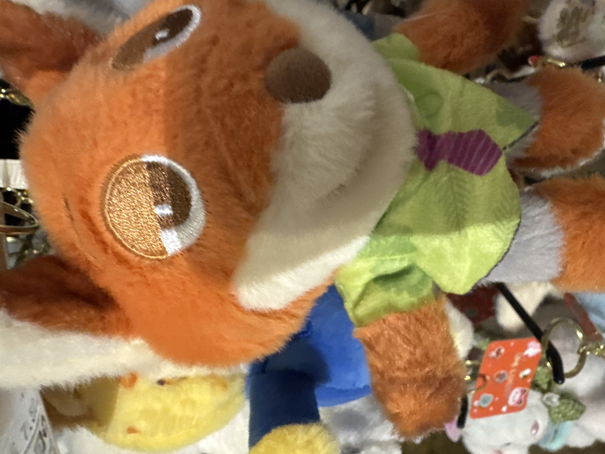 The image displays a close-up of a plush toy, prominently featuring its head and upper body. The main subject is an anthropomorphic animal, possibly a fox or similar creature, characterized by its bright orange fur, a white snout, and large, round eyes. The eyes are embroidered with a metallic, light brown sheen and distinct white highlights, suggesting a reflective quality. A small, dark brown circular nose is positioned on the white snout.
The plush toy is depicted wearing clothing: a light green shirt or vest with a prominent purple striped pattern on the front, and blue pants. Its fluffy orange arms and legs are visible, with white accents on the hands/paws. The background is blurred and appears cluttered, suggesting an indoor setting filled with other items. Partially visible below the main toy is a yellow plush object, and in the bottom right corner, several small charms or keychains can be seen, including one with an orange rectangular tag that has unreadable text and faint graphics. Other indistinct plush items, some white, are also scattered in the background. The overall scene suggests a collection of toys, possibly in a store display or a personal collection. There is no specific activity taking place; the image is a static portrayal of the plush toy.