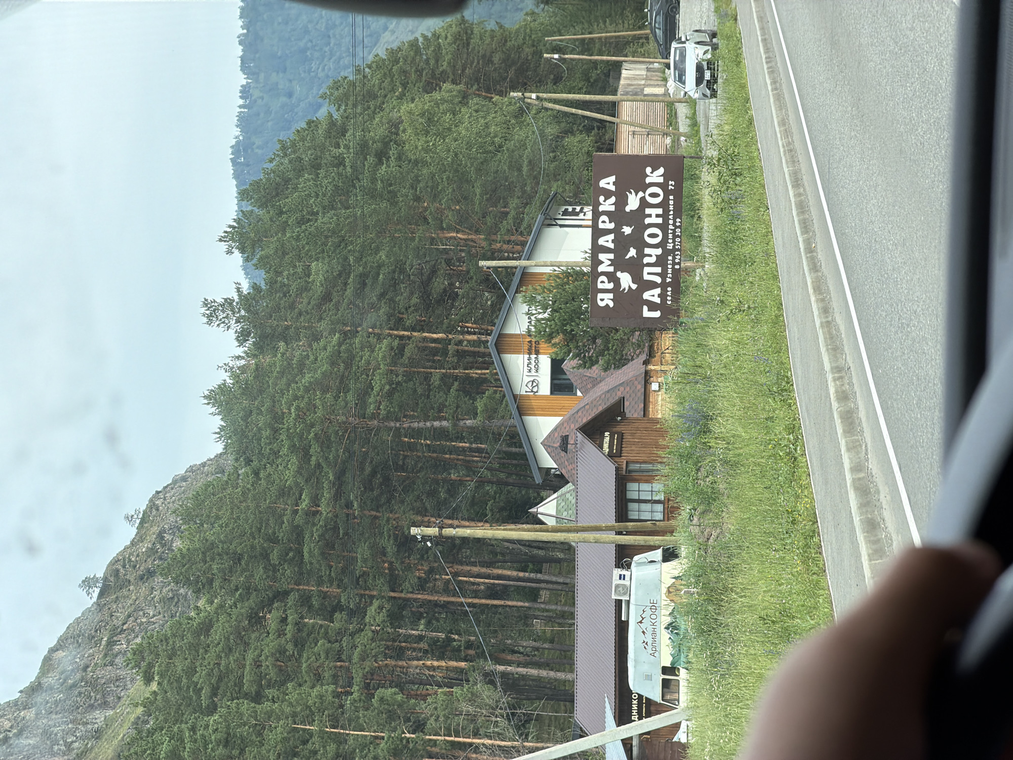 A brown, rectangular sign with white text is visible on the right side of the image, placed in a grassy area. The sign reads "ЯРМАРКА ГАЛЧОНОК" which translates to "FAIR GALCHONOK". Below this, it lists "село Узнезя, Центральная 73" and a phone number "8 963 570 30 99". A white car is parked to the right of the sign. In the background, there are several wooden buildings, a white van with "АРЛИАН КОФЕ" (ARLIAN COFFEE) written on it, and a dense forest of pine trees extending up a rocky mountain slope. The scene appears to be along a road, indicated by the asphalt and curb in the foreground. The setting is the city of Uznezya, Russia.