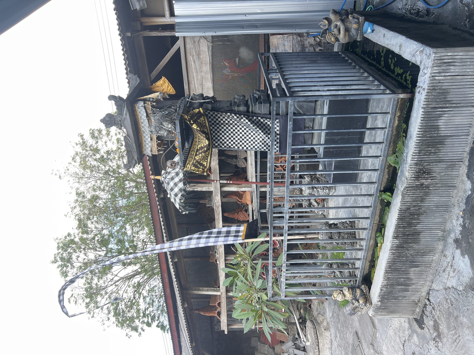The image depicts a Balinese temple-like structure, likely a small shrine or altar, situated outdoors in what appears to be a workshop or storage area. The central focus is a dark, intricately carved stone monument topped with a tiered roof adorned with gold and checkered fabrics. A tall, striped Balinese flag (kain poleng) curves gracefully from a pole to the left of the shrine.
Surrounding the shrine is a metal railing, behind which several small, carved stone figures are visible, some appearing to be statues of deities or guardians. In front of the railing, a low concrete structure contains a shallow pool of water with lily pads. Two small, seated figures are placed on either side of this pool, adding to the decorative or devotional aspect of the scene.
In the background, a long, open-sided structure houses shelves filled with what seem to be ceramic or clay art pieces, possibly decorative tiles or architectural elements, some arranged in rows. A green leafy plant grows to the left of the shrine, contributing a touch of nature to the scene. The overall environment suggests a place where religious artifacts or art are created or stored. The presence of traditional Balinese religious elements like the temple structure, the flag, and the statues strongly indicates a location in Bali, Indonesia. Given the artistic and artisanal context, it's plausible this scene is located within a craftsman's village or a workshop area in or around the city of Denpasar, known for its artistic heritage.