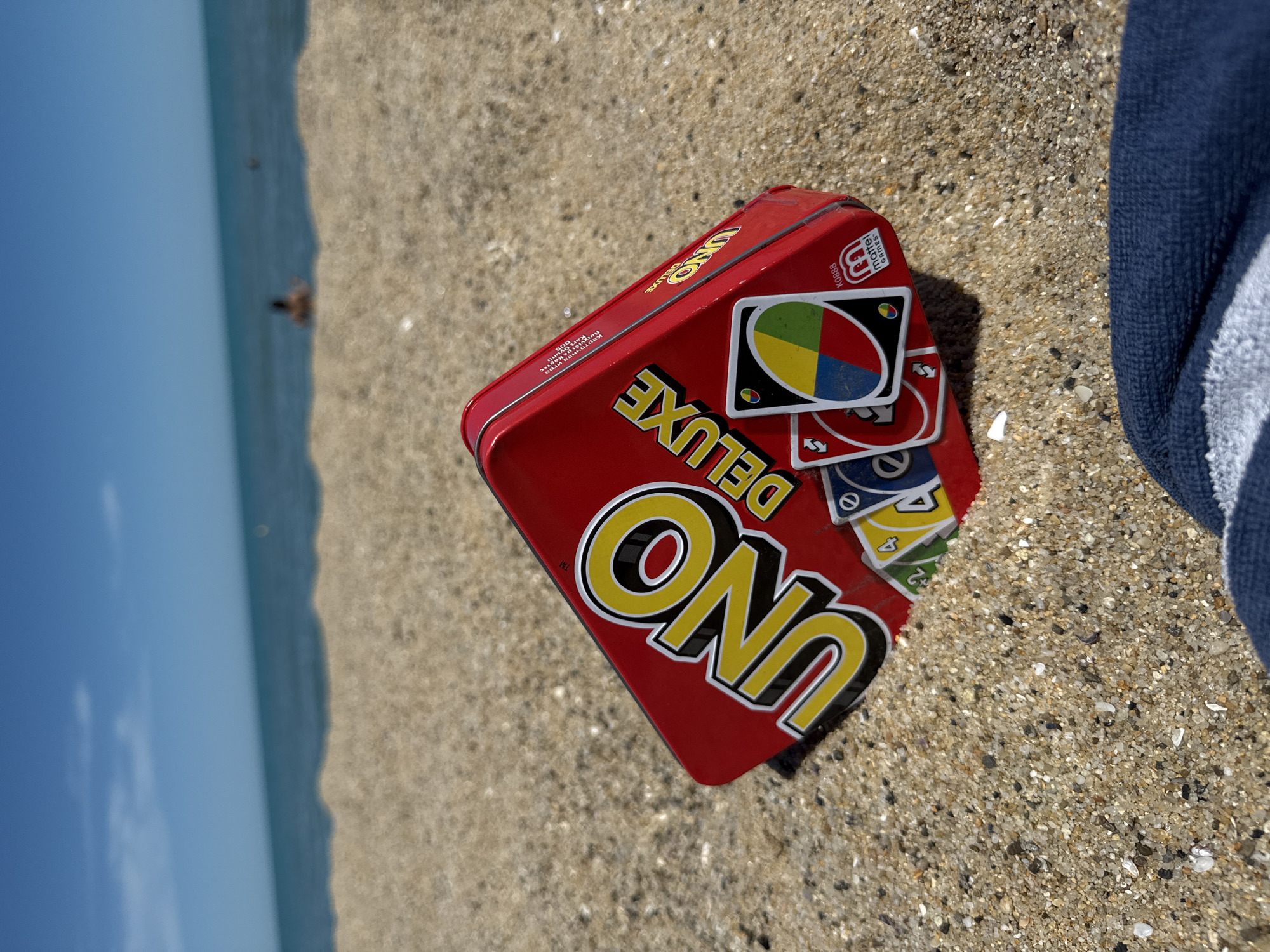 A red UNO Deluxe card game tin is placed on the sand at a beach, with several UNO cards visible underneath it. The sea and sky are in the background, with two blurry figures in the water. A portion of a dark blue towel is in the foreground. The scene appears to be at a beach in Varna, Bulgaria.