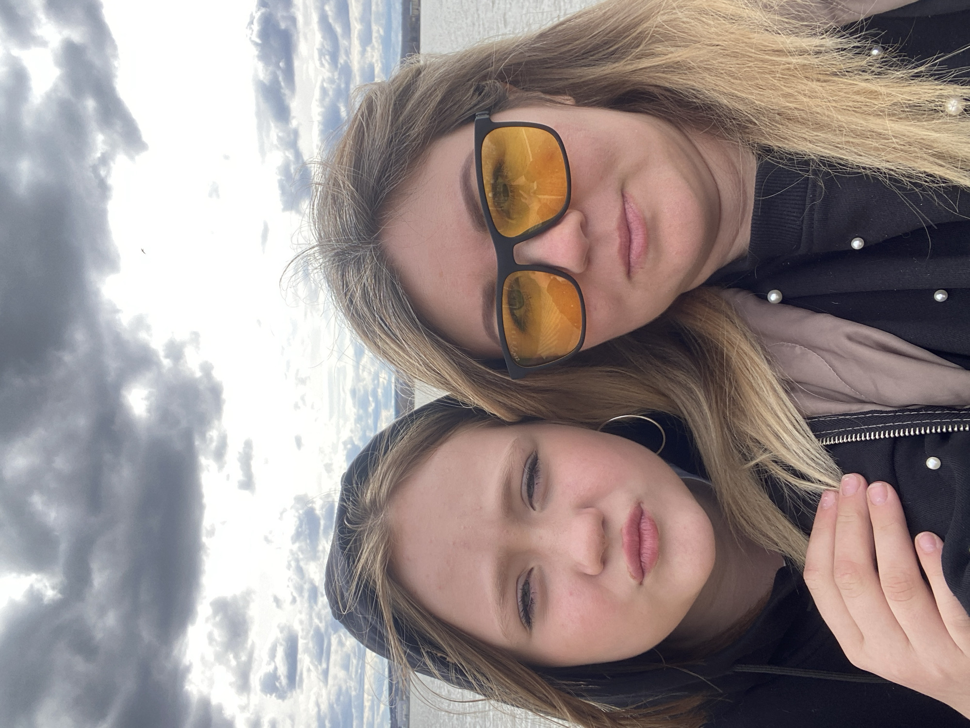 Two females, one older and one younger, are captured in a close-up selfie, filling most of the frame. The older female is positioned slightly behind the younger. She has long, straight blonde hair and is wearing black sunglasses with reflective orange lenses. Reflections in the lenses show a mix of sky and a faint, distant landscape or body of water. Faint text, including "BLIZZARD", is visible on the sunglass frame. She wears a black top adorned with small white pearl-like beads.
The younger female is in the foreground, looking directly at the camera with a neutral expression. She has long, light brown hair and wears a dark hooded top; a silver hoop earring is visible on her left ear. Her hands are resting near her chest, partially obscured by her clothing.
The background reveals an outdoor setting under a partly cloudy to overcast sky, with patches of brighter light visible amidst darker cloud formations. A small, dark bird is flying in the upper left portion of the sky. A subtle horizon line is visible in the distance, suggesting a large open space, possibly near water. The scene takes place during the daytime, and the clothing suggests a cool weather environment.