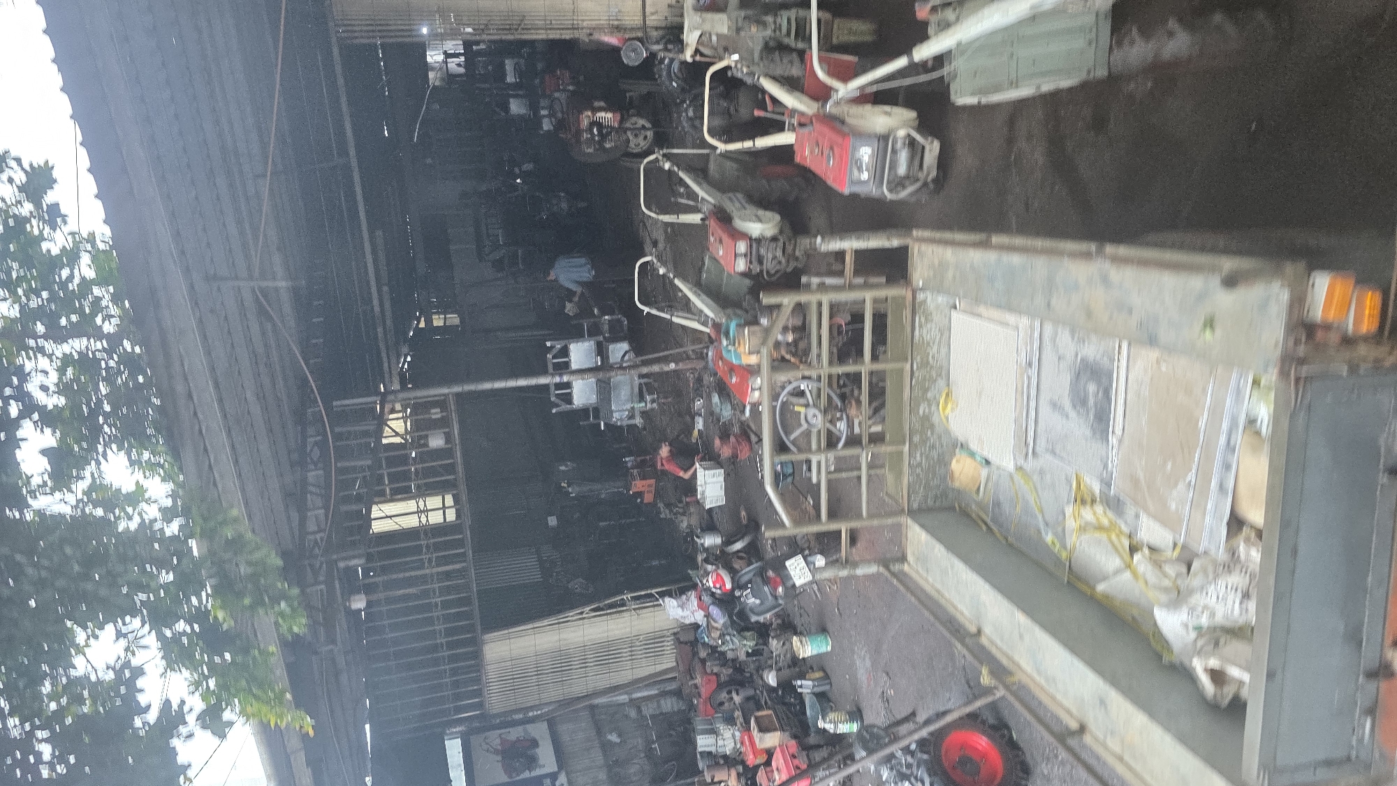 The image depicts a cluttered scene inside what appears to be a vehicle repair shop or storage facility in Pơng Drang, Vietnam. The location is identifiable by the license plates on the motorbikes. Various mechanical equipment, including several small tillers or cultivators, are visible. Some of the machinery is located inside the building, while other units are being loaded onto the back of a truck. The floor and surrounding areas are dirty and appear to be wet, suggesting a recent cleaning or rain. The building has a corrugated metal roof and siding, and there is foliage from nearby trees visible on the left side of the frame.