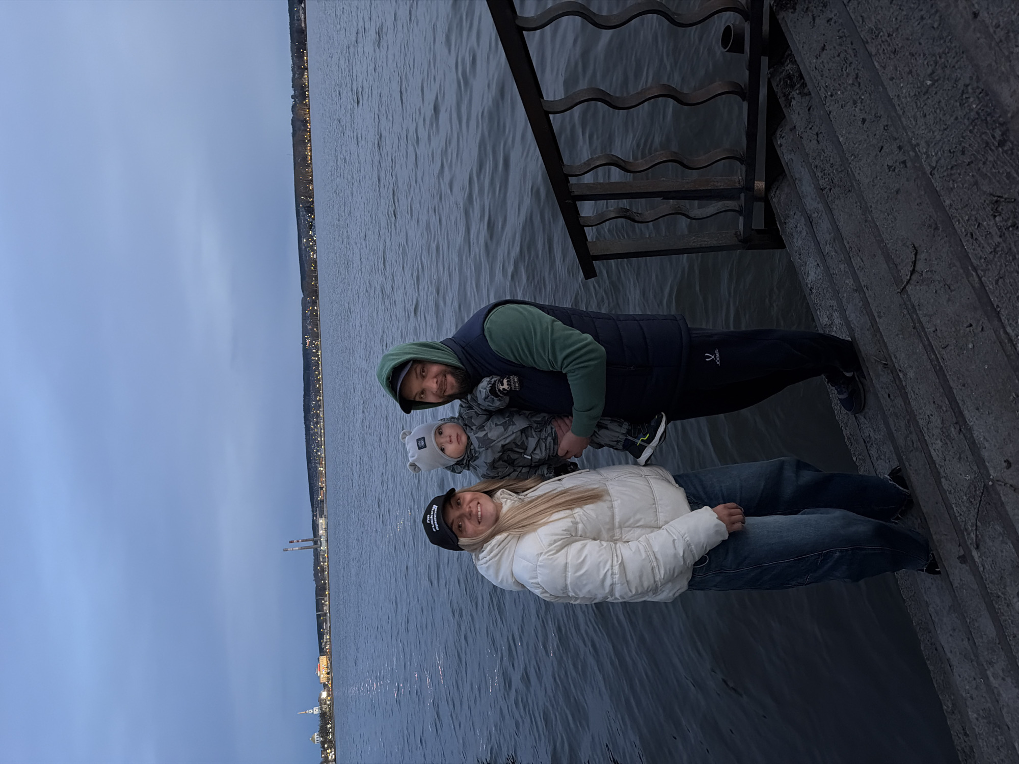 A family consisting of a man, a woman, and a child is standing on concrete steps next to a body of water. The man, wearing a dark vest over a green hooded sweatshirt, holds the child who is dressed in a camouflage-patterned outfit with a light-colored hat. The woman, wearing a white puffer jacket, a black hat with white lettering, and jeans, stands next to them. A dark metal railing is visible above the family, separating the steps from the water. The city of Votkinsk, Russia, is dimly lit in the background across the water. The sky is overcast.