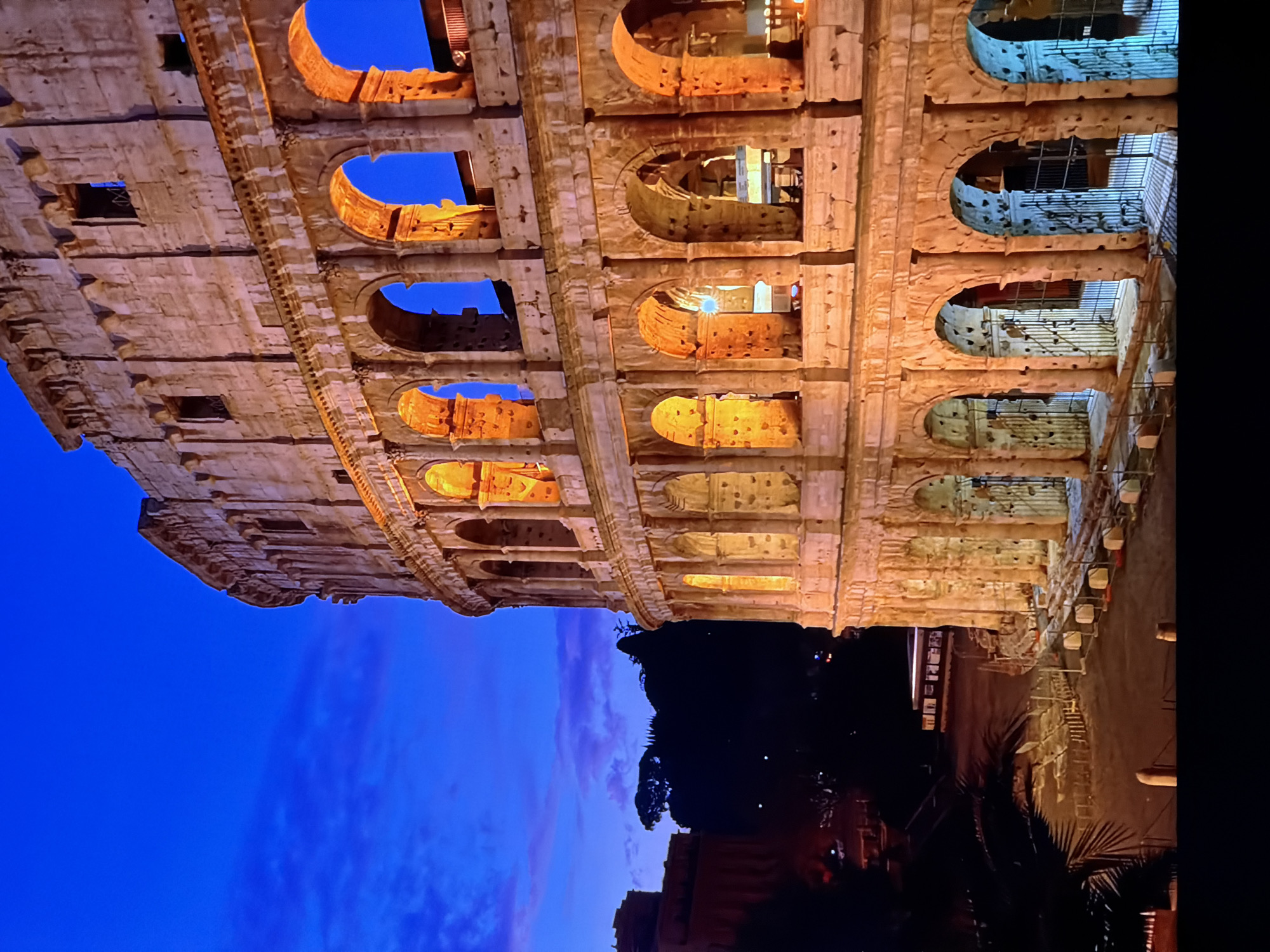 The image shows a portion of the Colosseum in Rome, Italy, at dusk. The ancient amphitheater is illuminated with warm, orange lights, highlighting its stone arches and weathered facade. The sky above is a deep blue, transitioning into darker hues, with faint traces of pinkish clouds. In the foreground, a pathway is visible with railings and some scattered fencing. The lower portion of the image shows a dark, blurred foreground, possibly indicating the perspective from a higher vantage point. The text provided stating the location is Chirchik, Uzbekistan is incorrect, as the image clearly depicts the Colosseum in Rome, Italy. There are no people visible in the image.
