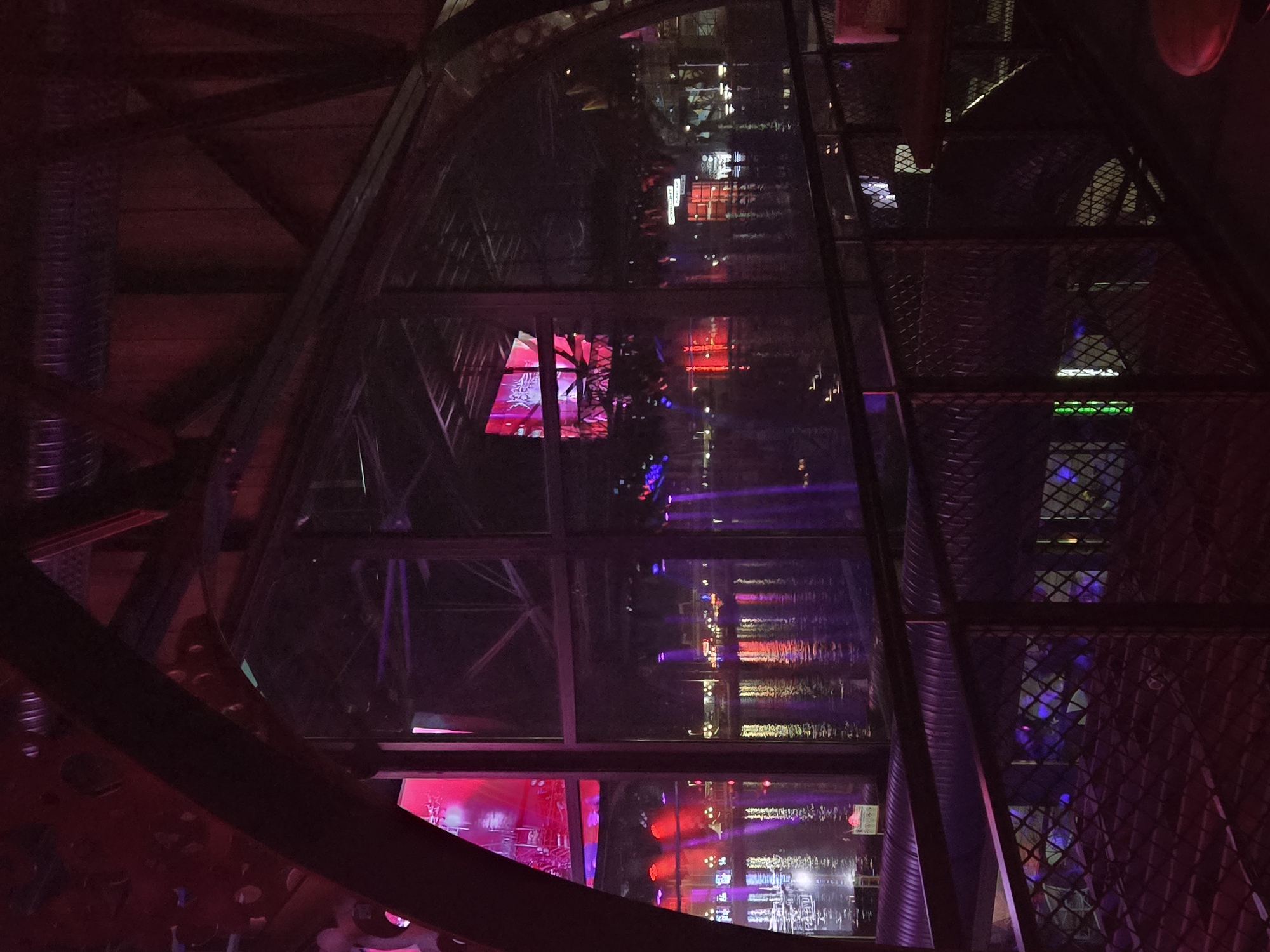 The image shows the interior of the Varna TV Tower in Varna, Bulgaria. The view is looking down the central shaft of the tower, with glass panels visible on either side. The tower's steel frame is visible, painted in a dark red or maroon color. Light reflections are visible on the glass panels, showing colorful lights and possible reflections from a cityscape outside the tower. The tower's interior is illuminated with red, pink, and purple lights. On the right side, there is a caged or mesh-like structure that forms the wall or side of the shaft. The general ambiance is dark, with the colorful lights providing the main source of illumination.