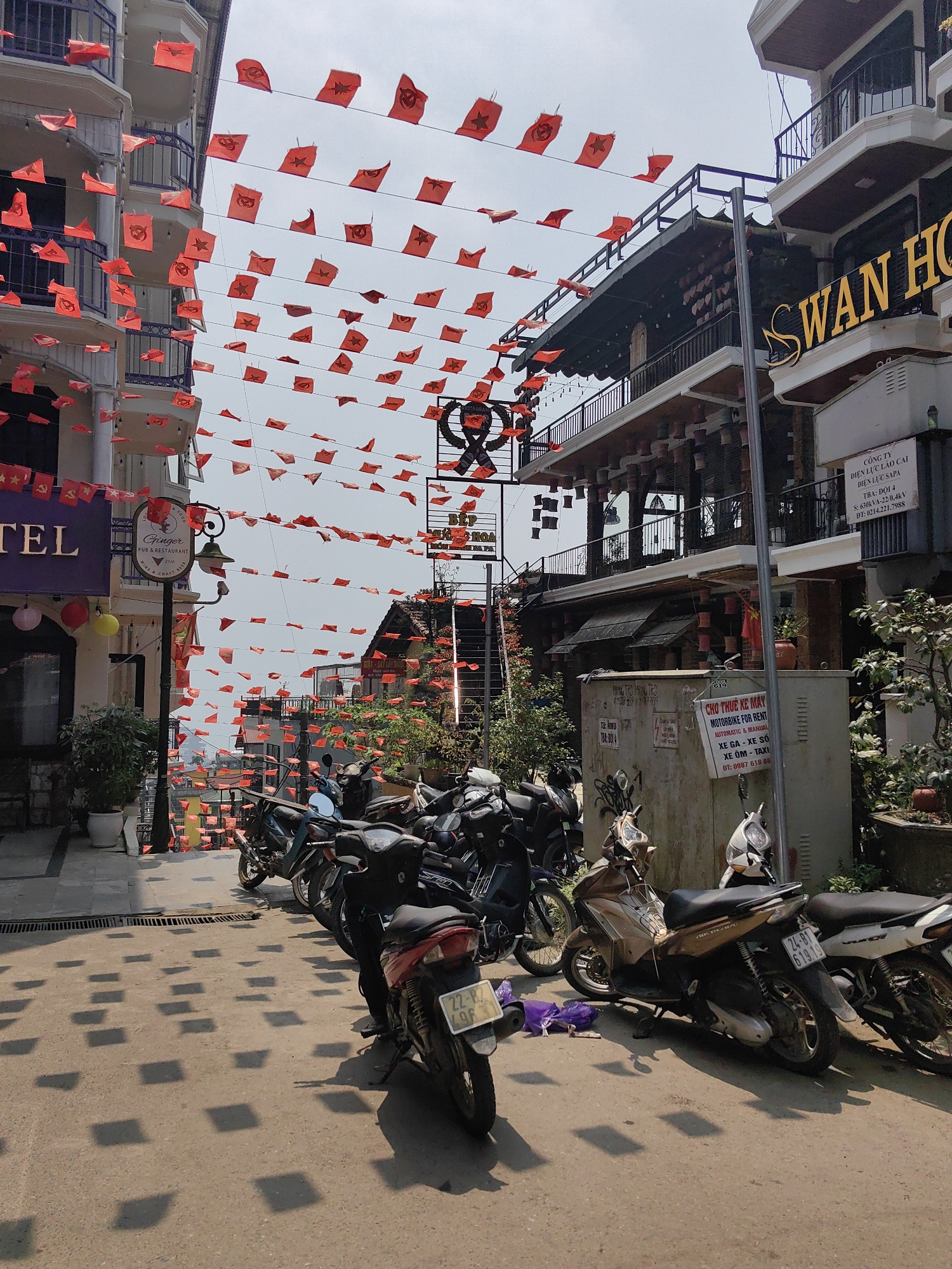 This forensic description details a daytime scene on a commercial street in Sa Pa, Vietnam.
The image captures a paved street flanked by multi-story buildings, extending uphill into the distance. Numerous small, red, rectangular flags, many featuring a yellow star and some a hammer and sickle emblem, are strung on multiple lines across the street overhead, casting distinct square shadows on the ground below. The sky above is bright white.
On the left, a white building with dark blue balcony railings has a partial purple sign reading "TEL," indicating a hotel. A decorative street lamp post in the foreground bears a sign for "Ginger Pub & Restaurant." Potted greenery is visible near the building's base.
On the right, a building displays "SWAN HO," identifying it as the Swan Hotel, with multiple balconies and a façade adorned with terracotta-colored pot-like decorations. Below this, a large, light-colored utility box covered in graffiti is positioned on the sidewalk. Attached to this box is a sign in Vietnamese and English, stating "CHO THUE KE MAY MOTORBIKE FOR RENT AUTOMATIC & MANUAL," providing details for scooter and taxi services. Above this, another sign mentions "CÔNG TY ĐIỆN LỰC LÀO CAI ĐIỆN LỰC SAPA," indicating an electricity company. Further down the street, another building with a darker facade is partially visible, with a sign containing the word "RESTAURANT." A ladder leans against a building in the middle distance on the right.
A significant number of motorbikes and scooters are parked in rows along the right side of the street and in the immediate foreground, facing generally uphill. Several scooters are clearly visible, with distinct features and license plates (e.g., "22-P2 496," "24-B1 619.19"). A purple plastic bag lies on the ground near one of the parked motorbikes.
No people are present or visible in the scene. The overall environment suggests a bustling commercial area, likely catering to tourism, given the hotel and restaurant signs and motorbike rental services.