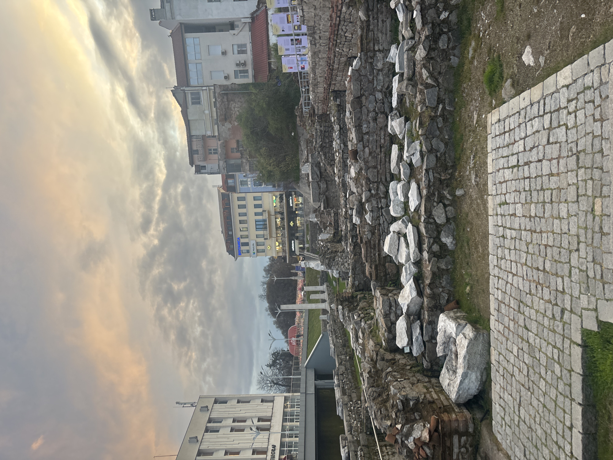 The image shows a section of the Ancient Stadium of Philippopolis in Plovdiv, Bulgaria. The ruins of the stadium are visible, composed of stone and brick. A paved walkway runs alongside the ruins. Buildings are visible in the background. The sky is cloudy, with sunset colors.