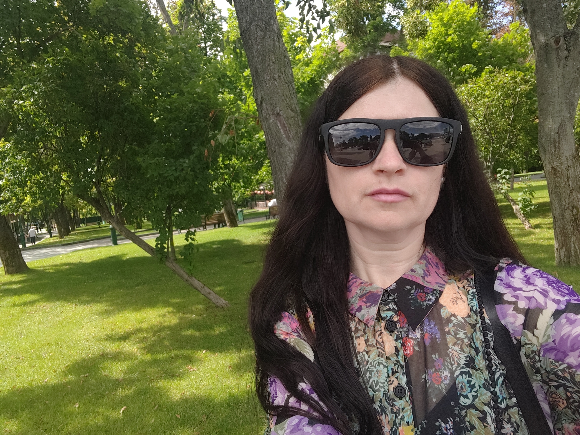 A woman with long, dark hair and sunglasses is taking a selfie in a park. She is wearing a colorful, floral print blouse with a black collar and black buttons. The background shows lush green grass, trees, and a paved pathway with a few people walking in the distance. Sunlight filters through the trees, casting shadows on the grass.