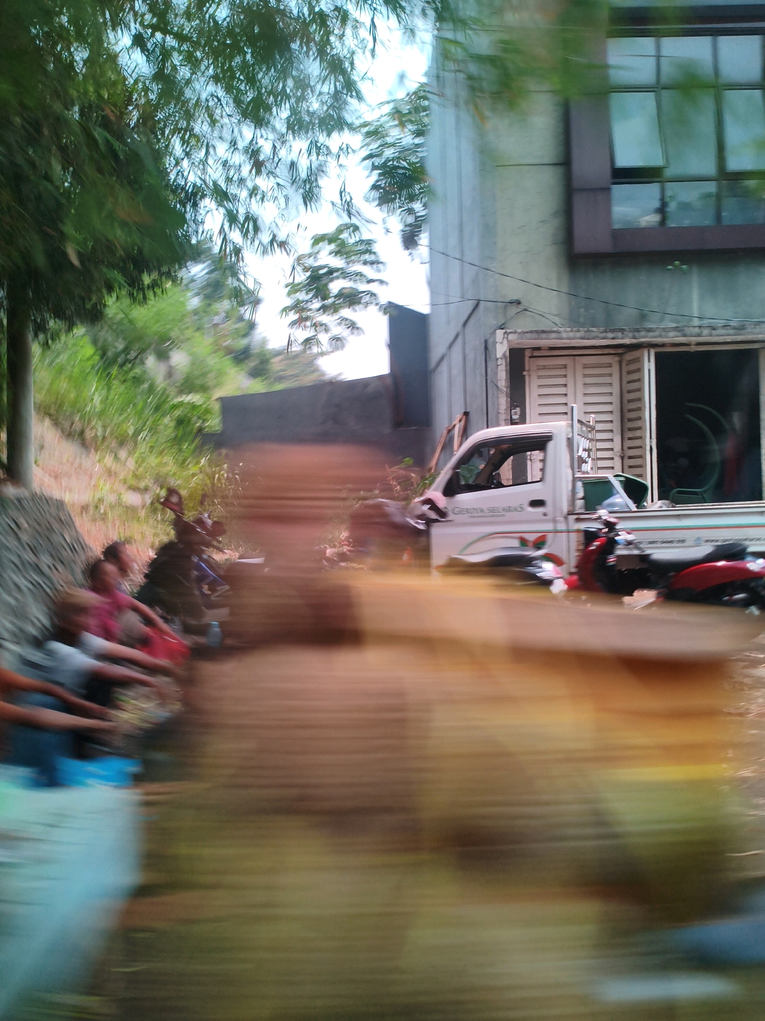 This is a motion-blurred photograph taken in Sukaraja, Indonesia, depicting a street scene with a truck and a motorcycle. In the foreground, several individuals, possibly locals, are seated or standing near what appears to be a motorcycle, with some blurry details of their clothing and skin tones. A bright blue object is visible to the left, and a textured, woven-like surface is present below the seated figures.
In the mid-ground, a white pickup truck is parked, with its doors and rear open. A red motorcycle is positioned in front of the truck. The truck has a white cab and a silver bed with green elements visible inside. The background shows a building with large windows, an area with lush green foliage, and a concrete structure. The overall impression is of a busy, perhaps informal, roadside area in Sukaraja, with elements of daily life captured in motion.