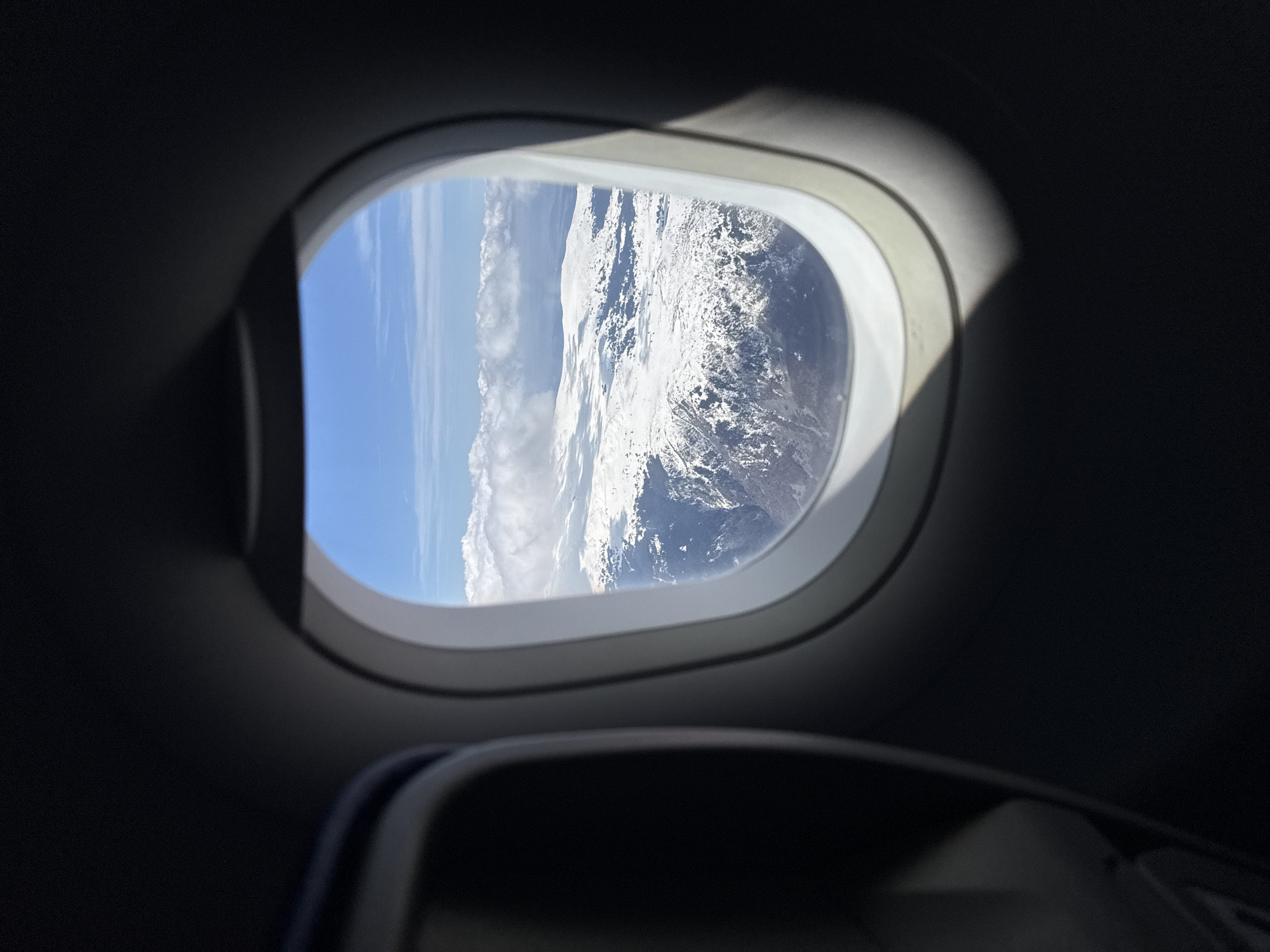 The image shows a view from an airplane window. Outside, there is a landscape of snow-capped mountains and clear blue sky. Clouds are visible. The interior of the plane is dark, with the window frame and the top of a seat visible in the foreground. This image doesn't have enough information to pinpoint a location in Busmantsi, Bulgaria.