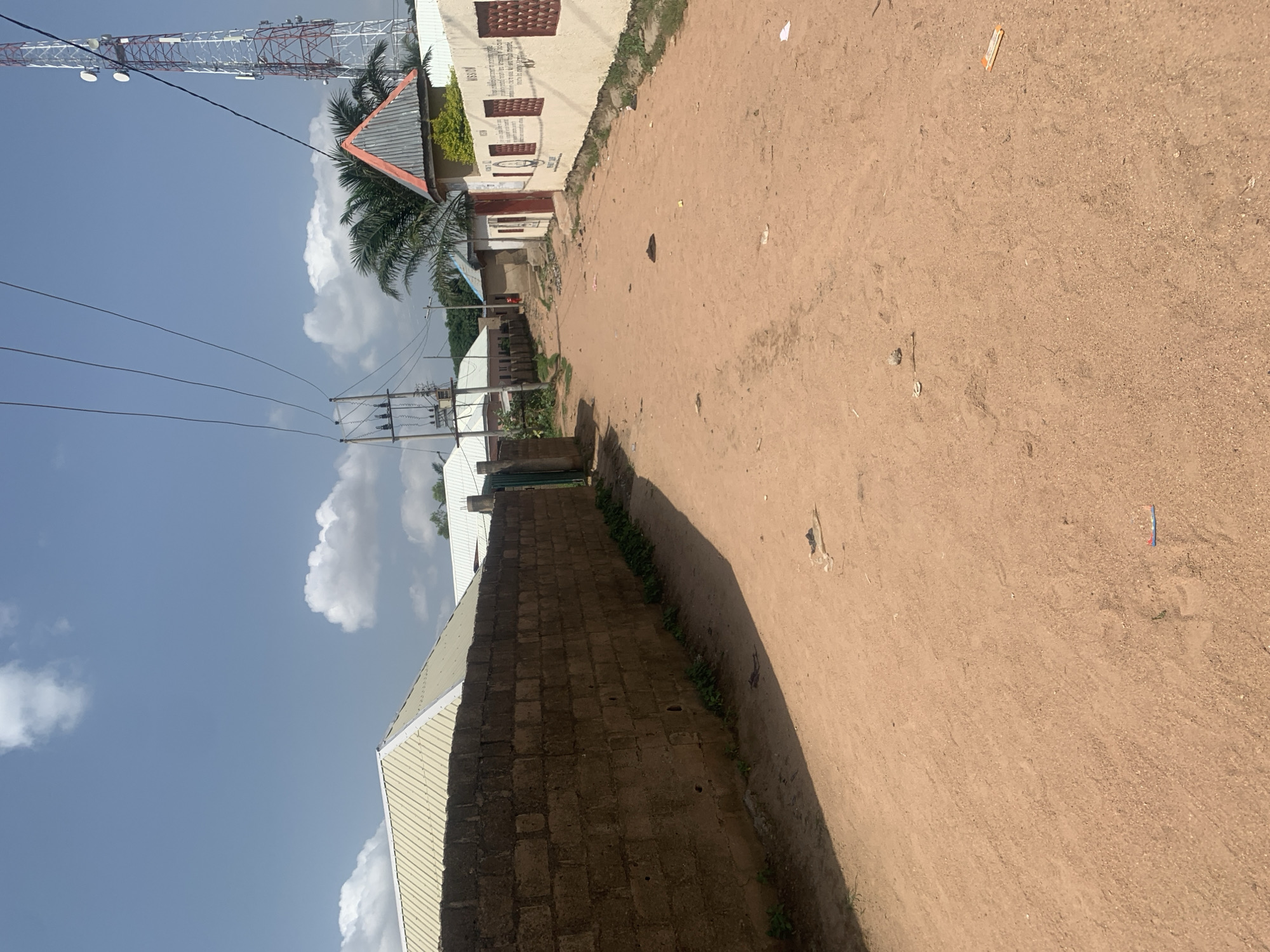 This is a photograph taken in Jalingo, Nigeria. The scene depicts a dirt road running alongside buildings and a communication tower against a bright blue sky with scattered white clouds. The road appears dry and dusty, with some scattered debris.
On the left side of the road, there are several buildings. One building has a light-colored facade with dark-colored window frames and some writing on the wall, which is partially obscured. There is also a building with a red-tiled roof and a large communication tower, possibly a cell tower, in the background, extending high into the sky. Power lines are visible strung between poles, indicating infrastructure in the area. Further down the road, more buildings with corrugated metal roofs are visible, suggesting a residential or commercial area.
The time of day appears to be daytime, likely morning or afternoon, given the bright sunlight casting shadows. The weather seems clear and sunny. The overall impression is of a scene in a developing urban or semi-urban area, characterized by its unpaved roads and functional architecture. No people are visible in the image. The writing on the building is too indistinct to be read.