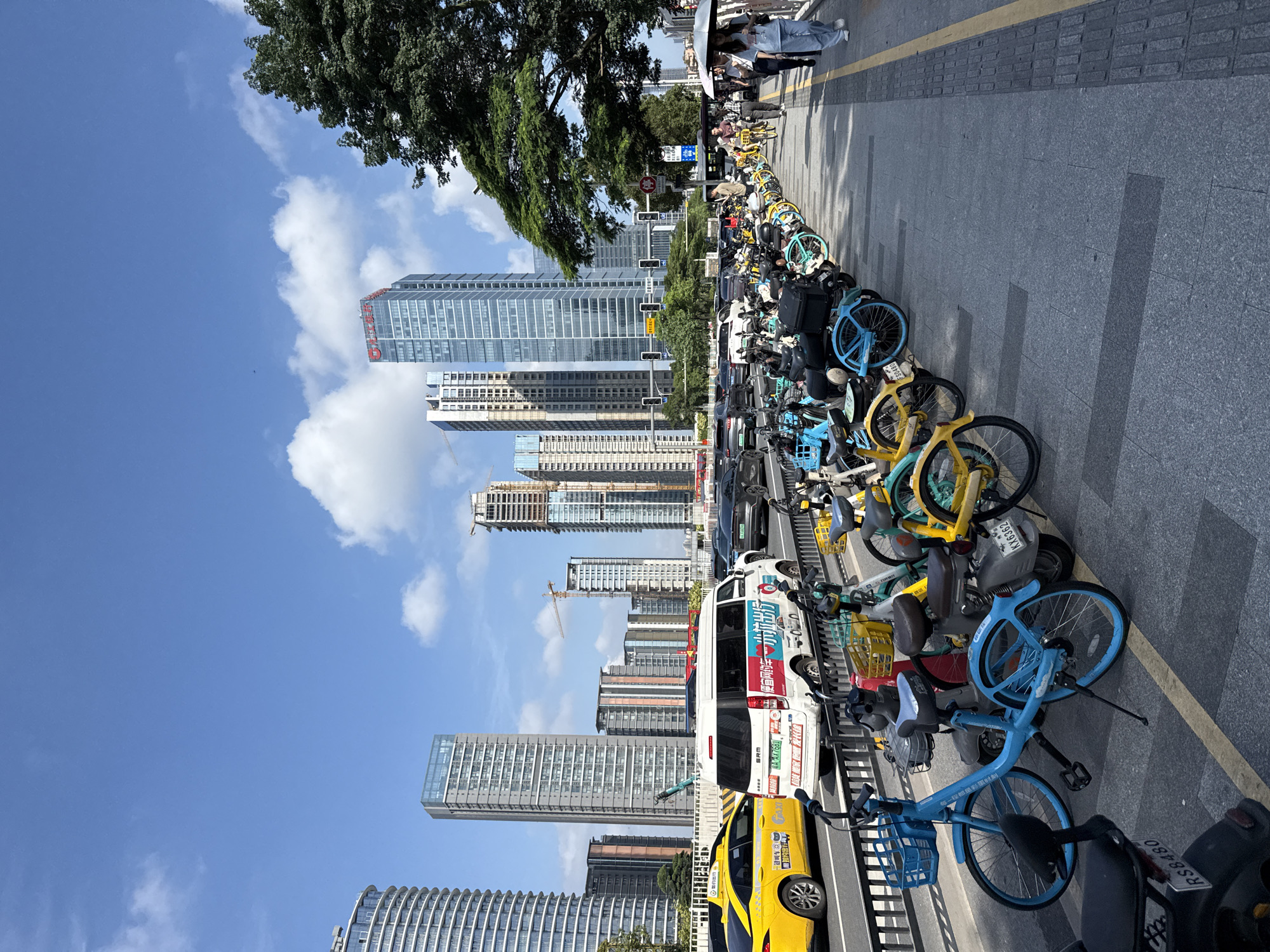 The image shows a street scene in Tianhe District, China, with a focus on a row of shared bicycles parked on a sidewalk. In the background, several modern skyscrapers dominate the skyline under a clear blue sky with scattered white clouds. A busy street runs parallel to the sidewalk, with a yellow taxi, a white van, and several other cars driving in traffic. On the sidewalk, a group of people are walking, some carrying umbrellas. The foreground features a line of colorful shared bicycles of various models and colors, including blue, yellow, and green, parked neatly in a designated area. Some of the bicycles have baskets and are equipped with kickstands. The street and sidewalk are paved with grey tiles. The overall scene depicts a typical urban environment with a blend of traffic, pedestrians, and a prominent presence of shared mobility options.