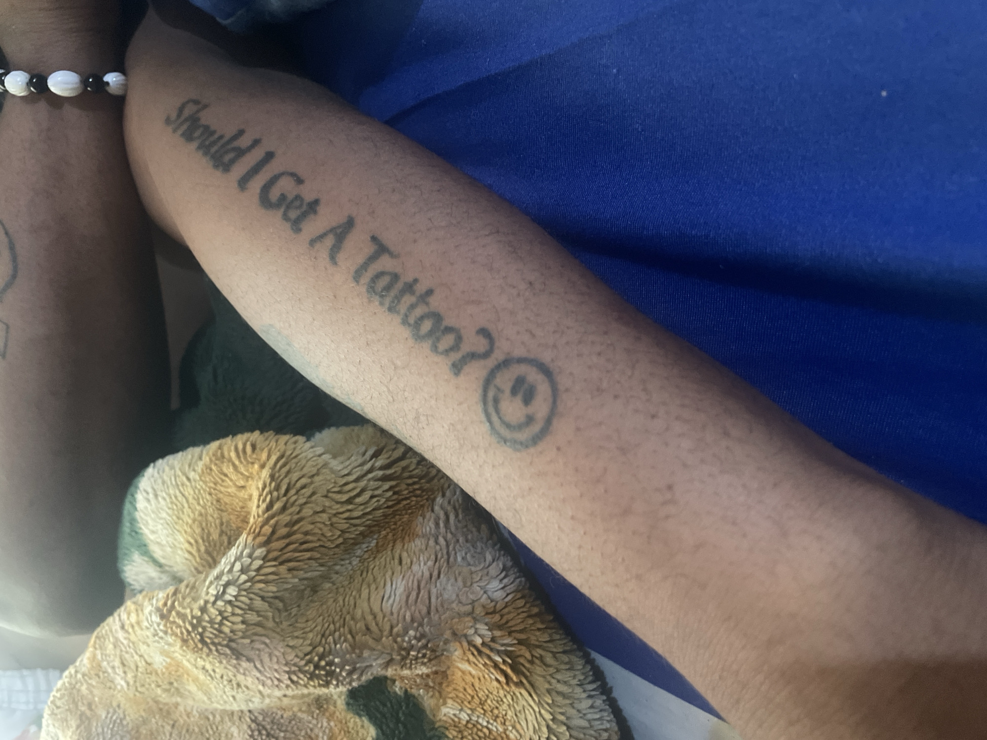 The image shows a person's arm with a tattoo that reads "Should I Get A Tattoo? :)" The arm is resting on a textured blanket and a blue surface. The image does not provide any information about the location in Jos, Nigeria, or any other details about the city or its inhabitants.