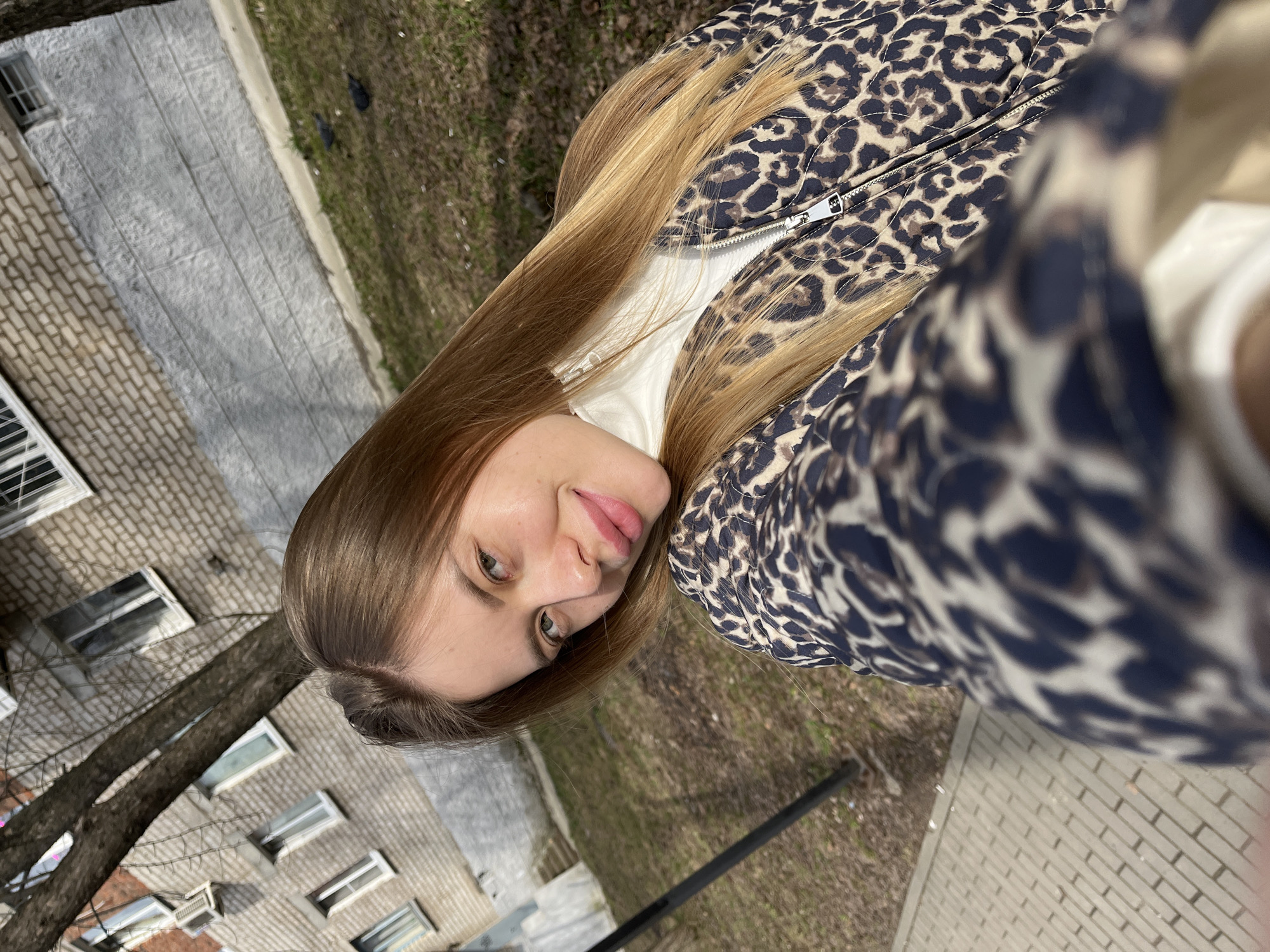 The image is a selfie of a young woman outdoors. She has long, light brown hair and green eyes, wearing a white turtleneck and a leopard-print jacket. The background suggests an urban environment, likely in the city of Chaikovsky, Russia. A brick building is visible on the left side of the frame with several windows. Adjacent to the building is a tree with bare branches. On the ground, there's a patch of grass with some bare spots, indicating possibly early spring or late fall. The bottom right corner shows a paved area with rectangular blocks, suggesting a walkway or sidewalk.