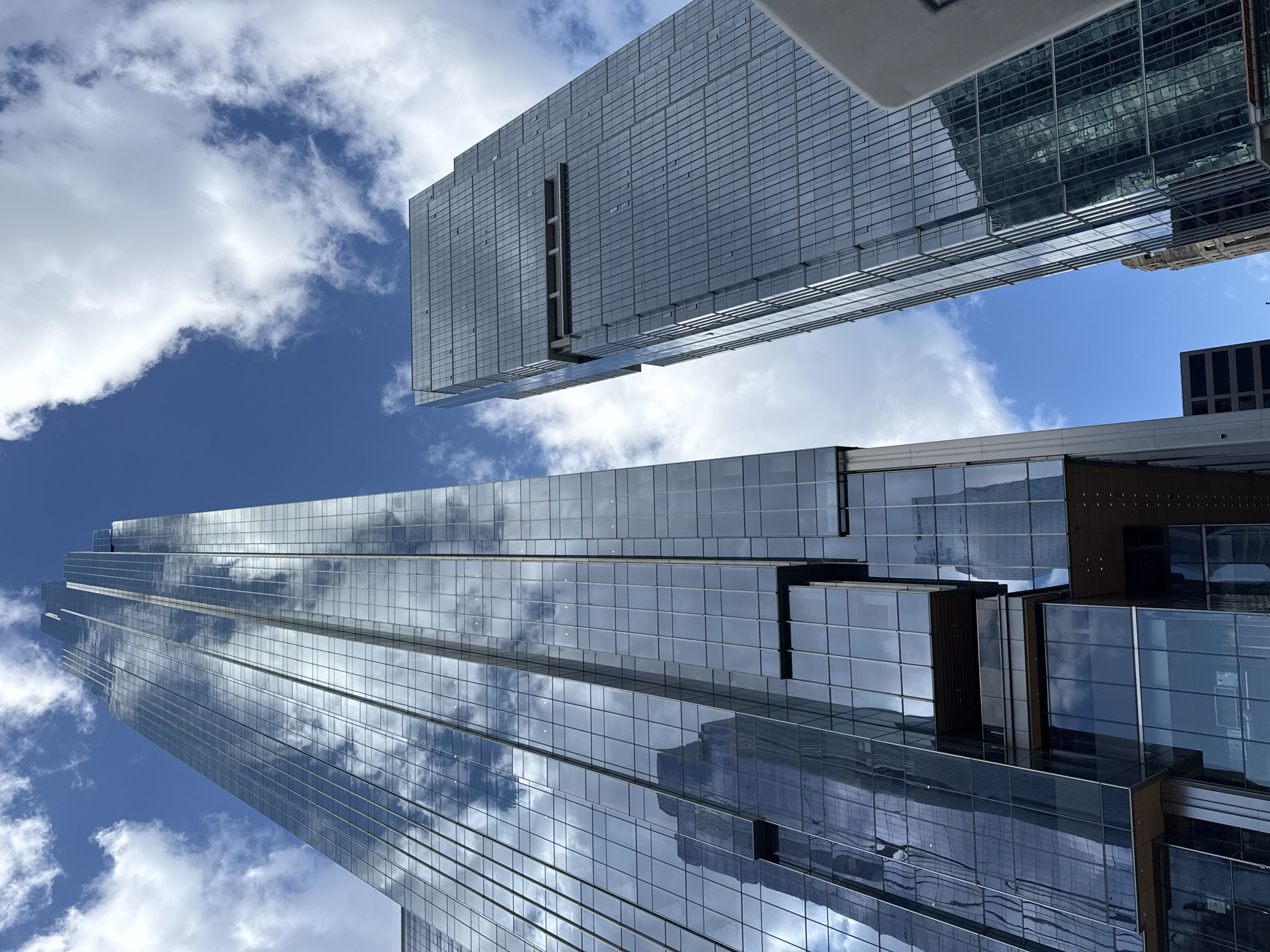 The photograph presents a low-angle view of modern architecture in Chicago, Illinois, USA. Two primary high-rise buildings dominate the frame, both characterized by glass facades reflecting the sky and clouds. The structure on the left appears taller and more slender, extending upwards at an angle, its surface filled with the reflected imagery of the blue sky and cumulus clouds. The building on the right is positioned at a sharper angle relative to the viewer, and also reflects the clouds, but is wider and less tall. A bright blue sky with scattered white clouds is visible in the negative space between the buildings. The lighting suggests a sunny day. The perspective is from street level, looking up towards the skyscrapers.