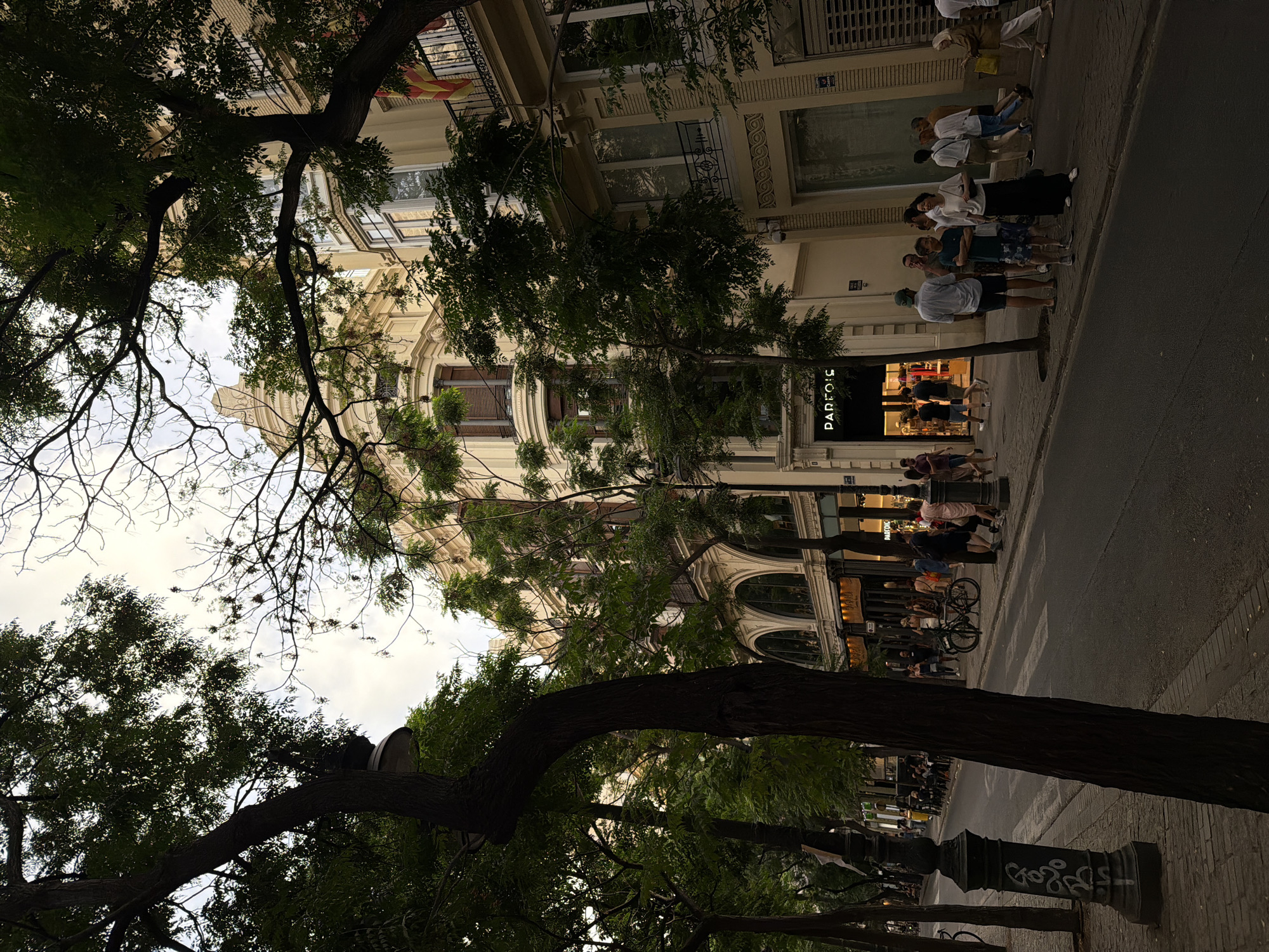 The image depicts a street scene in a city, likely in Europe, with a focus on the architecture and the people present. The buildings are multi-story structures with ornate detailing, featuring arched ground-floor storefronts and balconies with wrought-iron railings. One building displays a flag that appears to be a regional flag of Spain. Trees line the sidewalk, their branches casting shadows across the street.
Several people are visible on the sidewalk and crossing the street. They are casually dressed, suggesting a warm day. Some individuals are walking, others are standing in small groups, and a bicycle is parked near the curb. The storefronts include a shop with the illuminated sign "PARFOIS." The street itself is paved, with a pedestrian crossing marked by white stripes. The lighting suggests it is either late afternoon or early evening, with a diffused sky. The overall atmosphere is one of a lively urban environment.