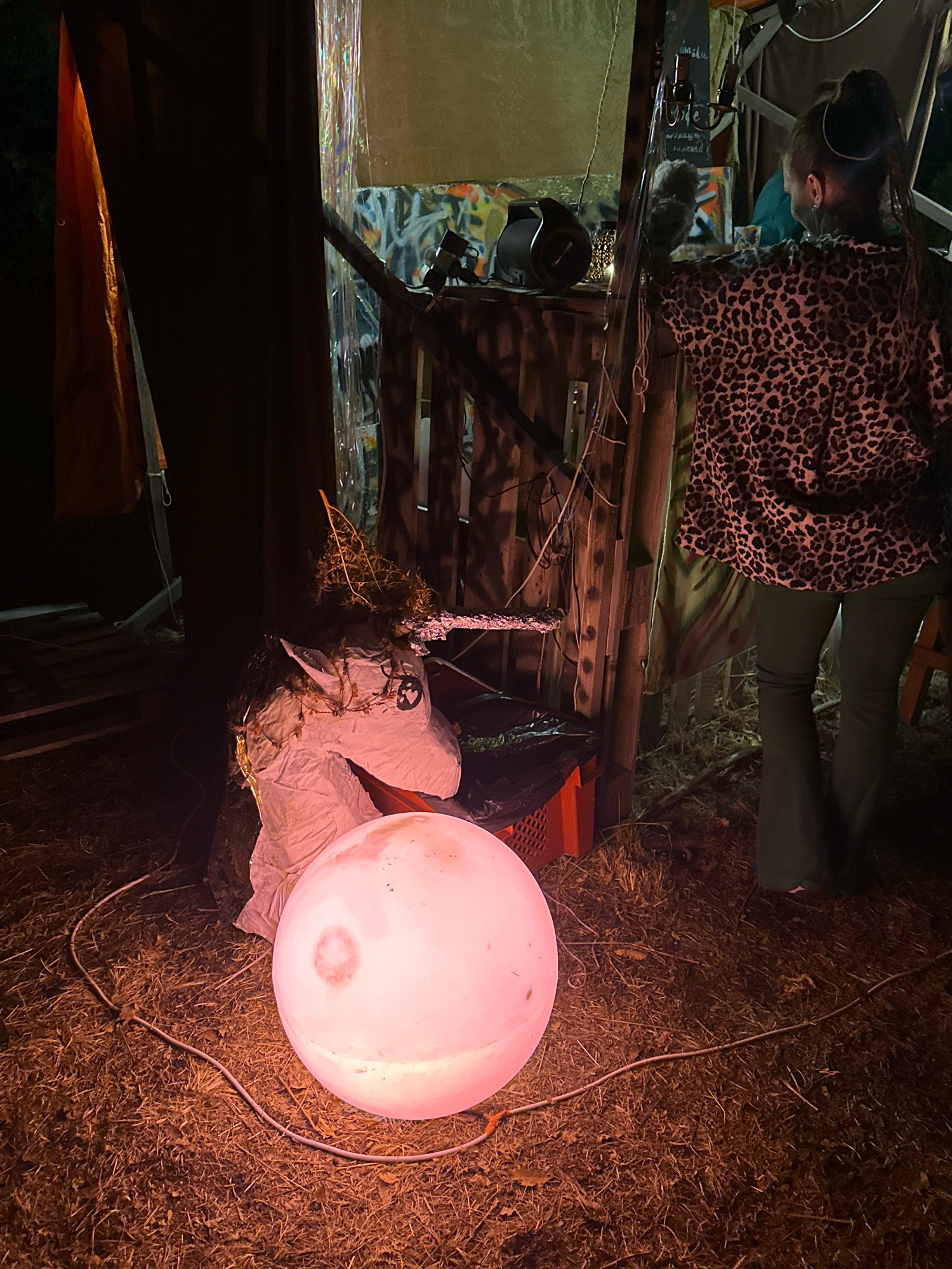 A large, pink illuminated sphere sits on dry grass in front of a makeshift structure. To the left of the sphere is a crudely made unicorn head, crafted from what appears to be paper mache and adorned with twigs and moss. Behind the unicorn, a red plastic crate is visible, containing a black plastic bag. The structure behind these objects is made of wood and corrugated metal, with graffiti art on the walls. A woman wearing a leopard-print top and dark green bell-bottom pants stands near the structure, facing away from the camera and towards the interior. The overall scene suggests an outdoor event or installation in Cetinje, Montenegro.