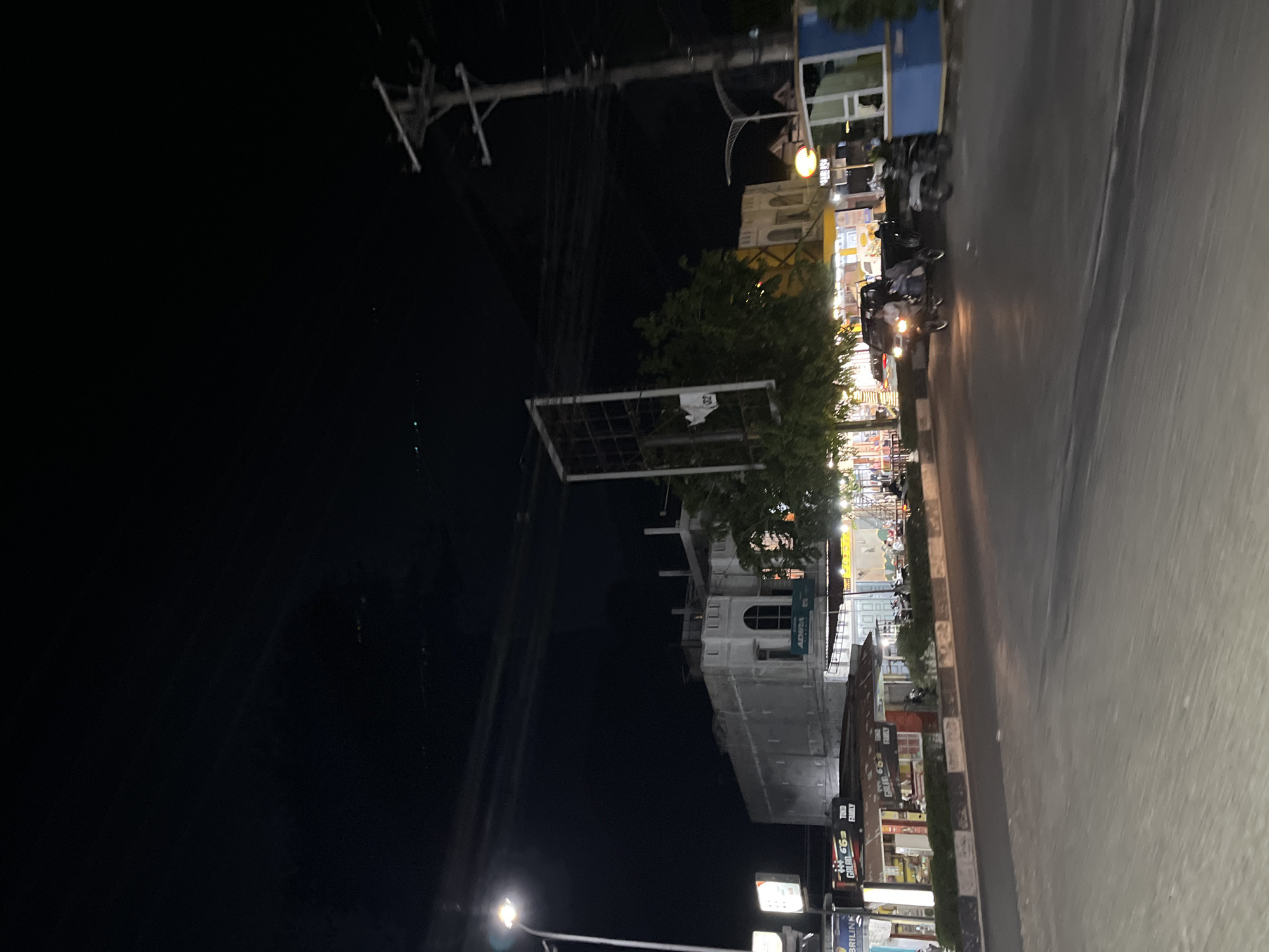 The image captures a nighttime street scene in Langsa, Indonesia. A streetlamp illuminates the foreground, casting light on a road with a distinct curb and median. Across the street, a row of buildings is visible, some with lit windows and signs. One building appears to be under construction or renovation, with a white facade and scaffolding. Various shops and stalls line the street, with some people visible in and around them.
A few vehicles are present, including a three-wheeled vehicle with two people on it, and a motorcycle parked on the side. There are also power lines and utility poles running overhead, common features of urban environments. The overall atmosphere is one of a typical bustling street at night, with the lighting creating a dynamic interplay of light and shadow. The presence of Indonesian signage and the architectural style of the buildings suggest the location is indeed within Indonesia, consistent with the provided location information.