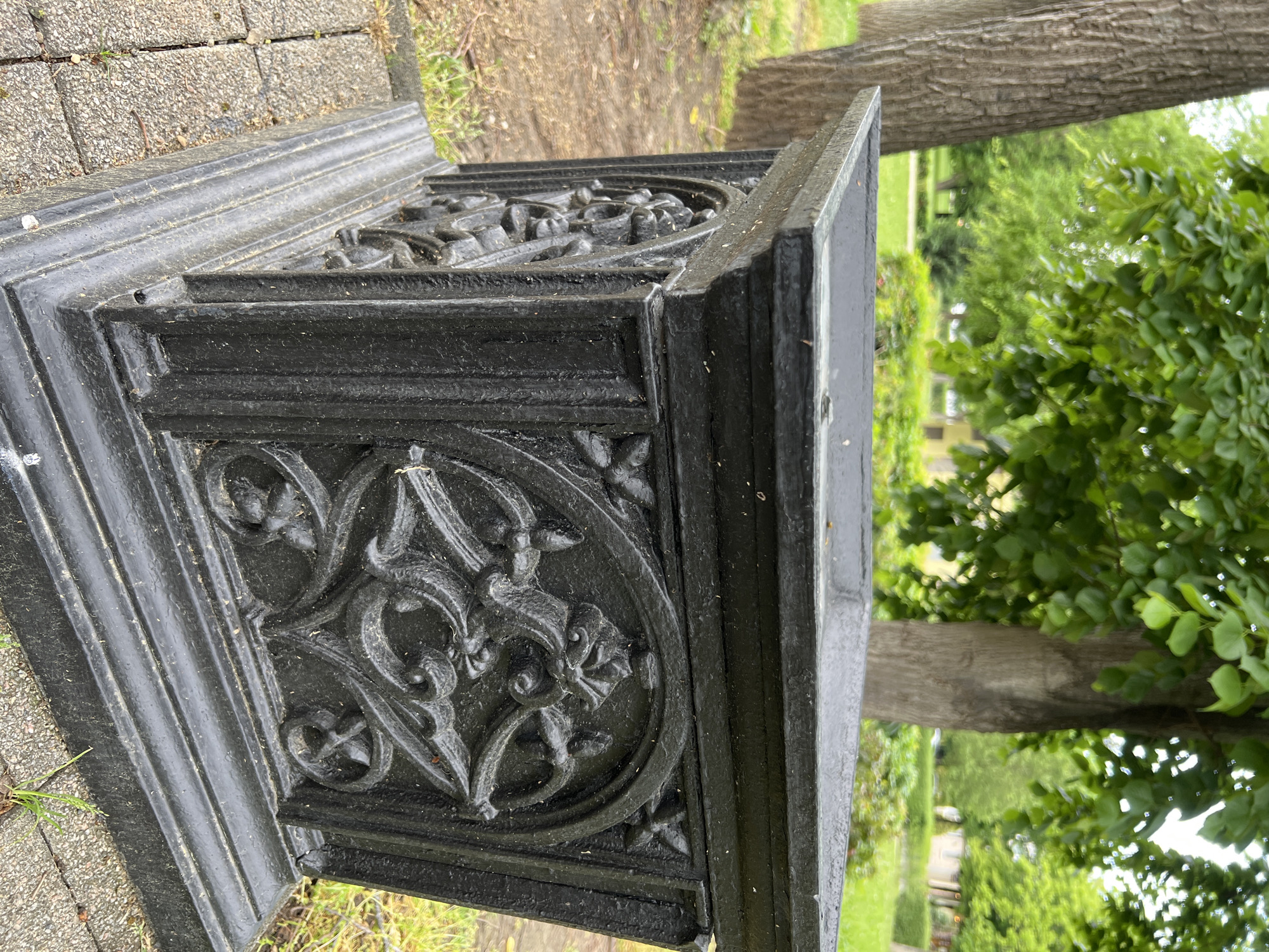 A low-angle, medium shot shows the corner of a black, decorative, cast-iron structure. The structure sits on a paved surface. An intricate design of circular and floral motifs is on the side. The background features grass, trees, and possibly other structures. The photograph appears to have been taken outdoors in a park-like environment. The location in the city of Krasnodar, Russia, is not evident from the image.
