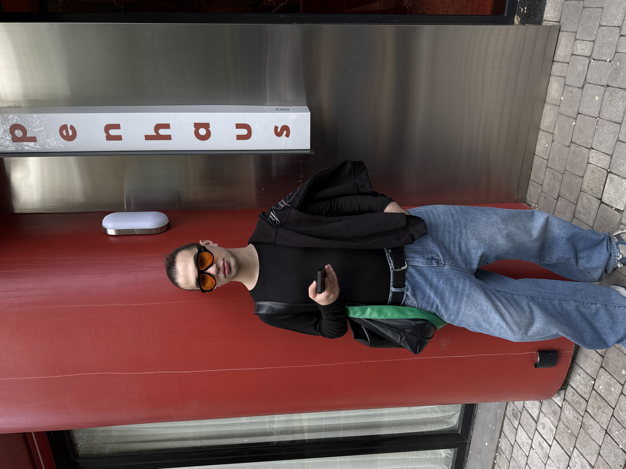 A full-length, medium shot depicts a man posing outdoors in Yerevan, Armenia. He wears orange sunglasses, a black long-sleeved shirt, light-wash jeans, and a black belt. He holds a smartphone in his right hand. A black jacket is draped over his left shoulder, and a black purse with a green strap hangs from his right shoulder. He stands in front of a red and silver building with the word "Supra" spelled out vertically on a white sign. The background includes a window, a stone sidewalk, and parts of the building's exterior.