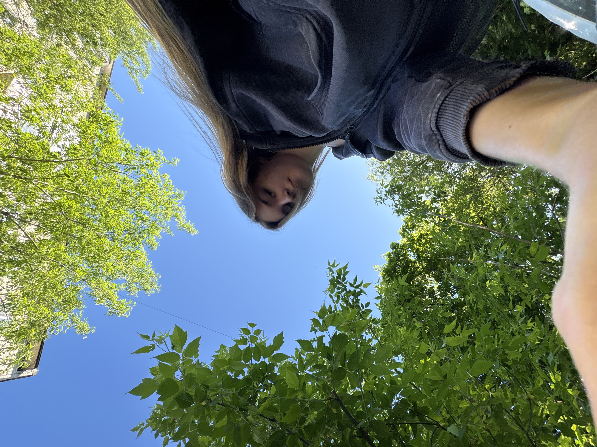 The image is a low-angle selfie taken outdoors in Perm, Russia. A fair-skinned person with long, light brown hair is looking down at the camera, wearing a dark-colored hoodie. The background consists of a bright blue sky and green foliage from trees. A building facade is partially visible in the upper left corner.