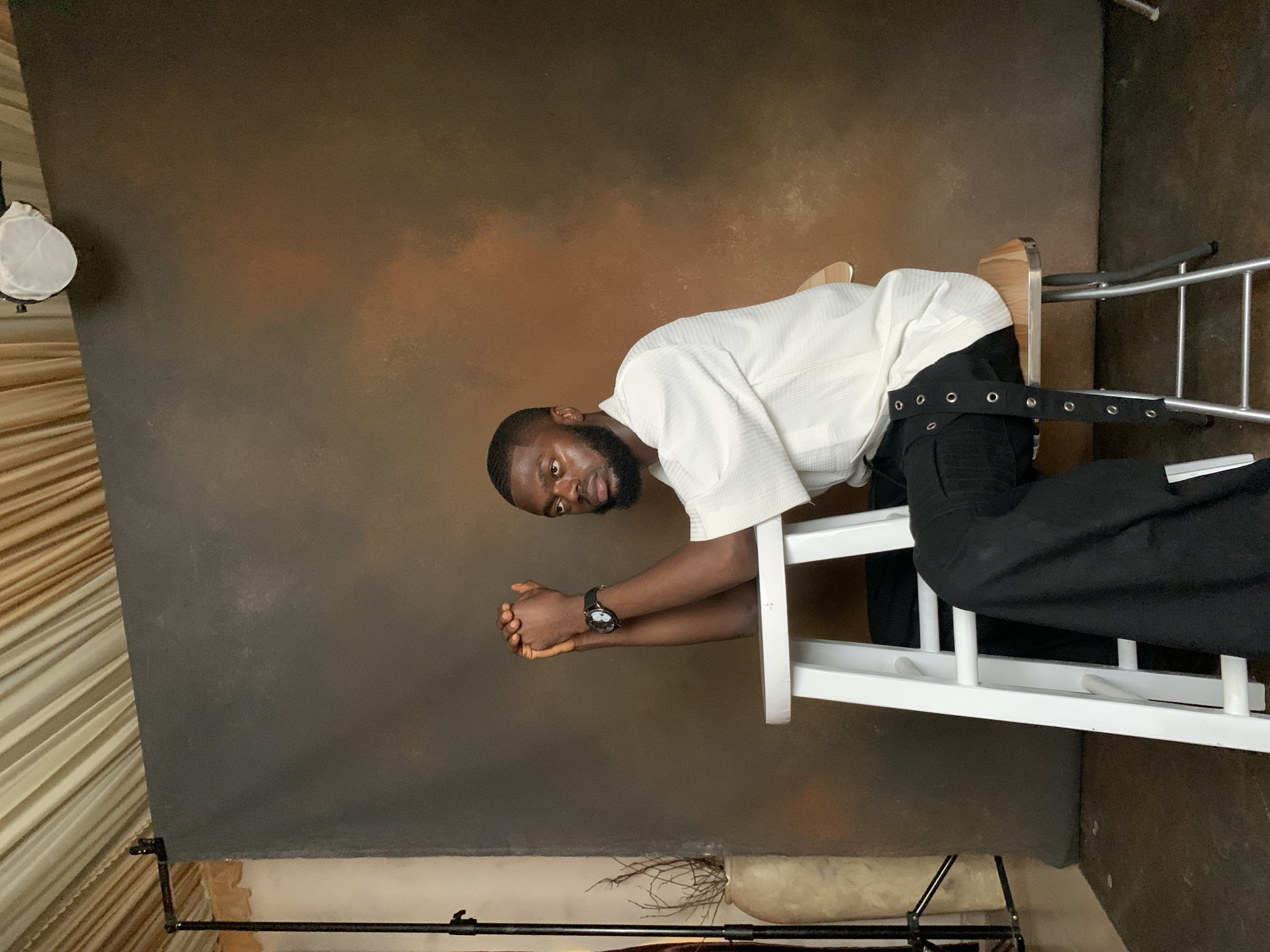 A young Black man with a beard and short hair is seated on a white stool. He is wearing a white, textured, short-sleeved shirt and black pants with buckle detailing. His hands are clasped in front of him, resting on his forearms. He is looking to his right. The background is a textured brown backdrop. To the left, a tall beige vase with dry branches is visible, and further left, a black light stand. The scene appears to be a photo studio. The location is Barakin Sabawa, Nigeria.