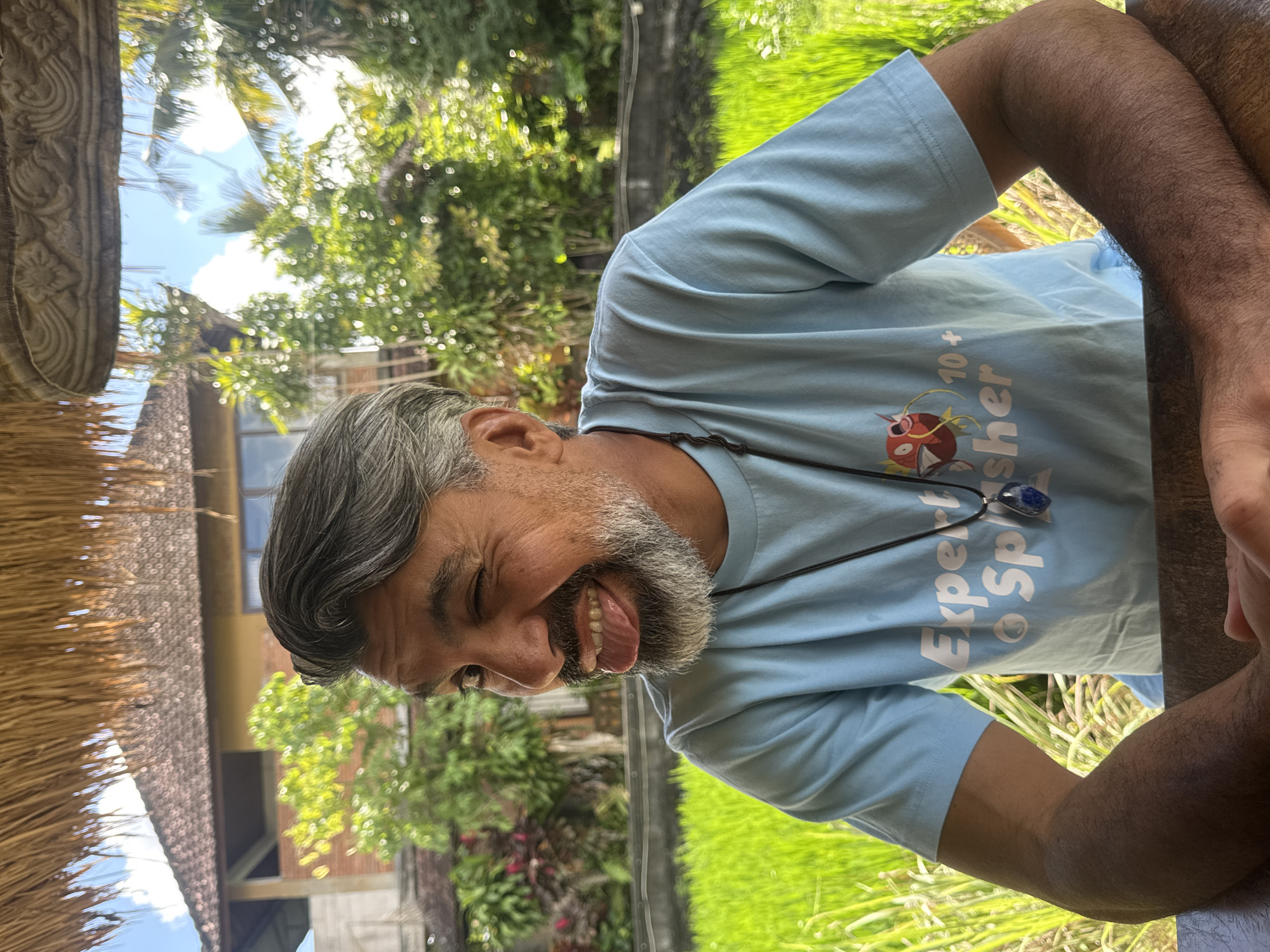 A man with grey hair and a grey beard is sitting at a wooden table, with his arms resting on it. He is wearing a light blue t-shirt with a graphic design on it and a necklace with a blue pendant. He is sticking his tongue out and winking. In the background, there is a lush green rice paddy, some tropical foliage, and a building with a tiled roof. The scene appears to be outdoors, under a thatched roof structure. The location is Tegallalang, Indonesia.