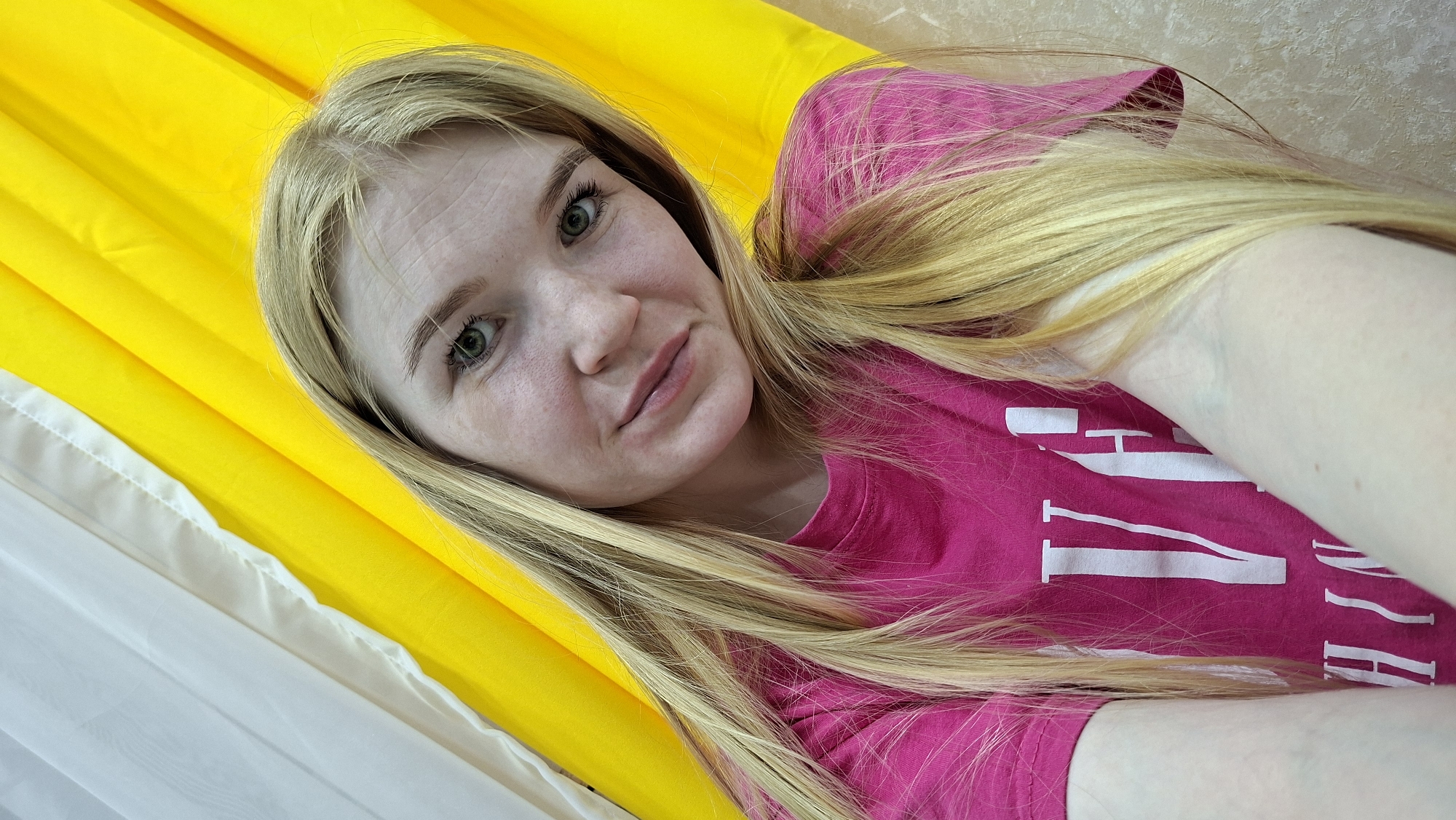 A fair-skinned woman with light blonde hair is positioned in front of a background consisting of white and yellow fabric, possibly a curtain or draped material. She is wearing a bright pink T-shirt with white lettering. The photo is a close-up, selfie-style shot capturing her face and upper body. The location is presumed to be in the city of Chaikovsky, Russia, based on unspecified provided context.